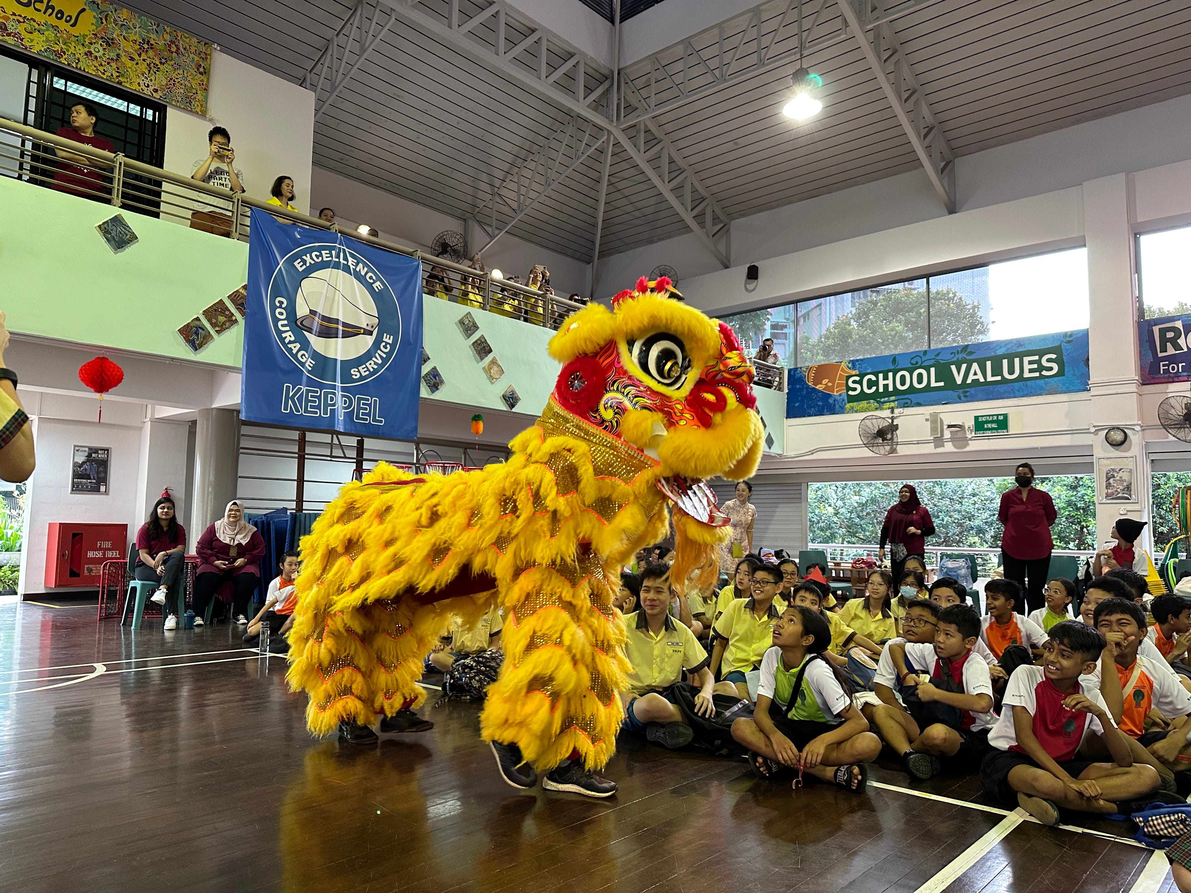 Yellow lion dance costume performs in a school hall as seated students watch, with banners on the walls.
