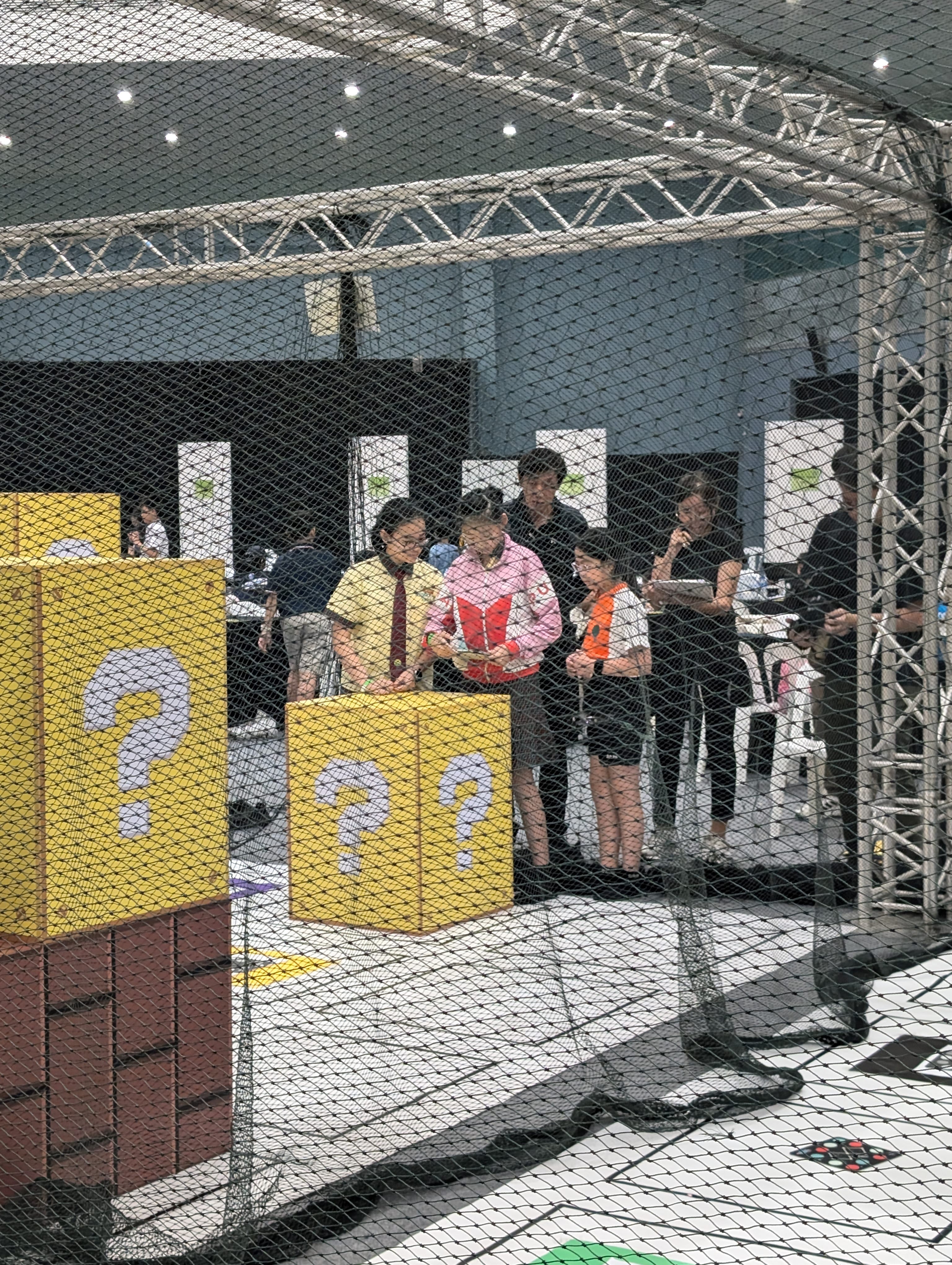 People in a netted booth at an expo, beside large yellow blocks with question marks.