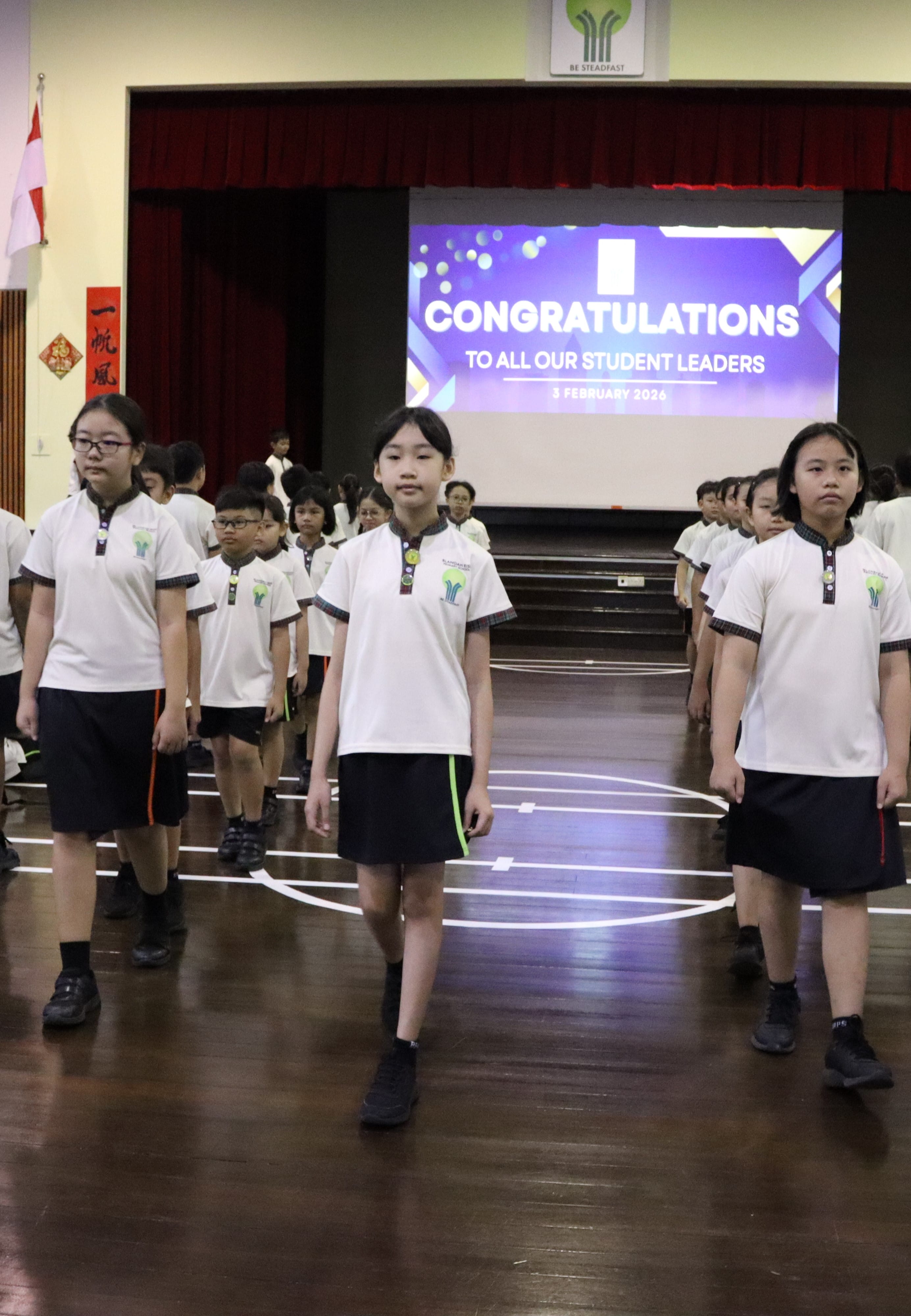 Students in white uniforms march in a school hall with a screen reading Congratulations to All Our Student Leaders.