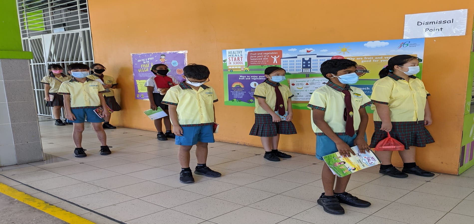 Children in yellow school uniforms and face masks line up at a school dismissal point near health posters.