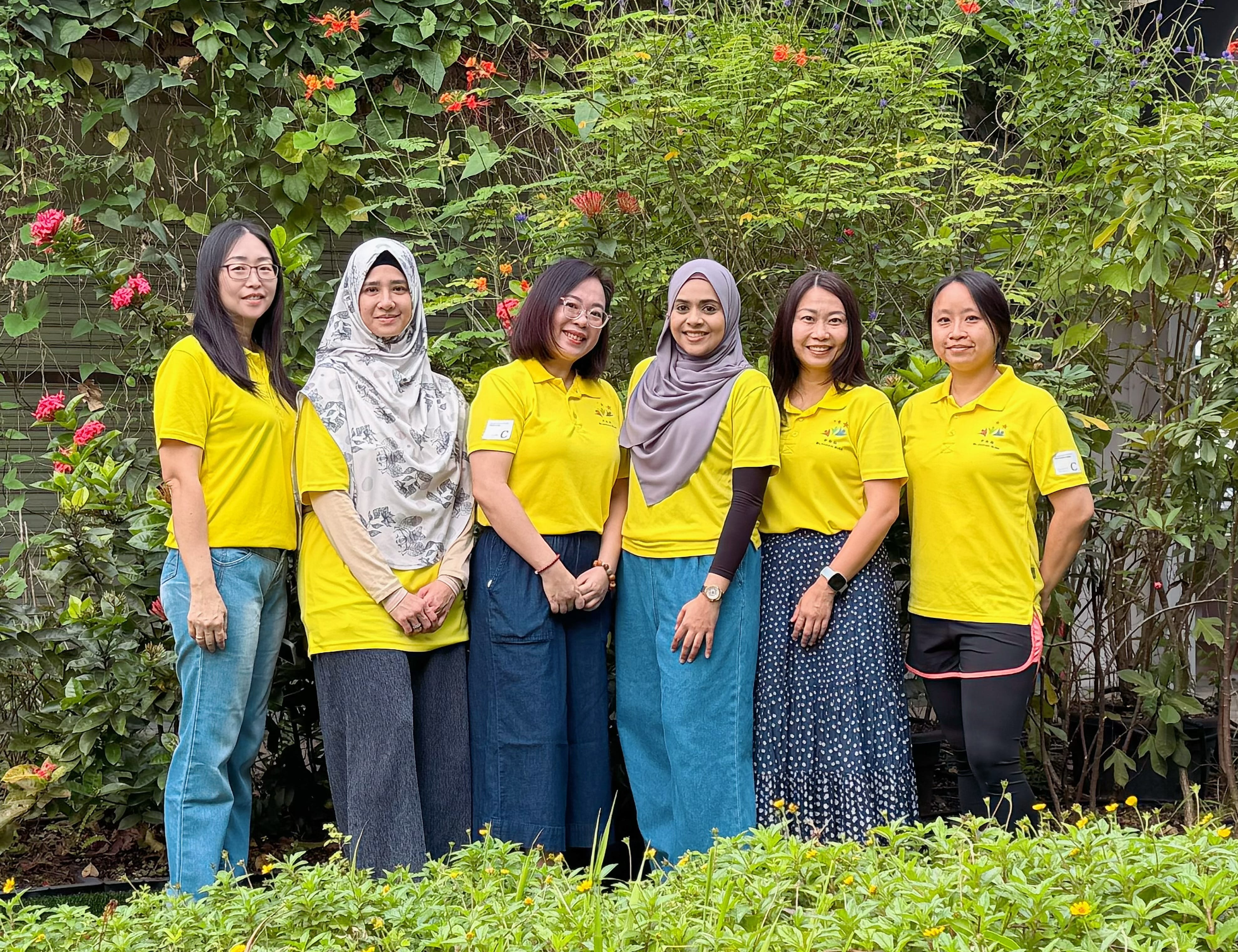 Six women in yellow shirts pose in a garden with green leaves and red flowers behind them.