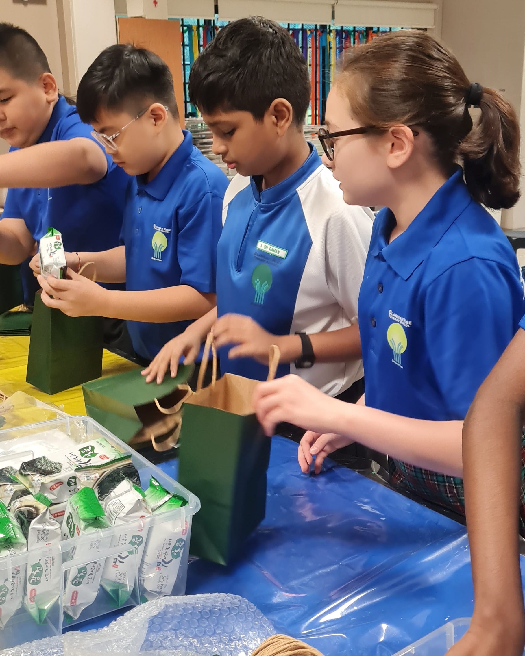 Four children in blue school uniforms pack items into green and brown paper bags at a table.