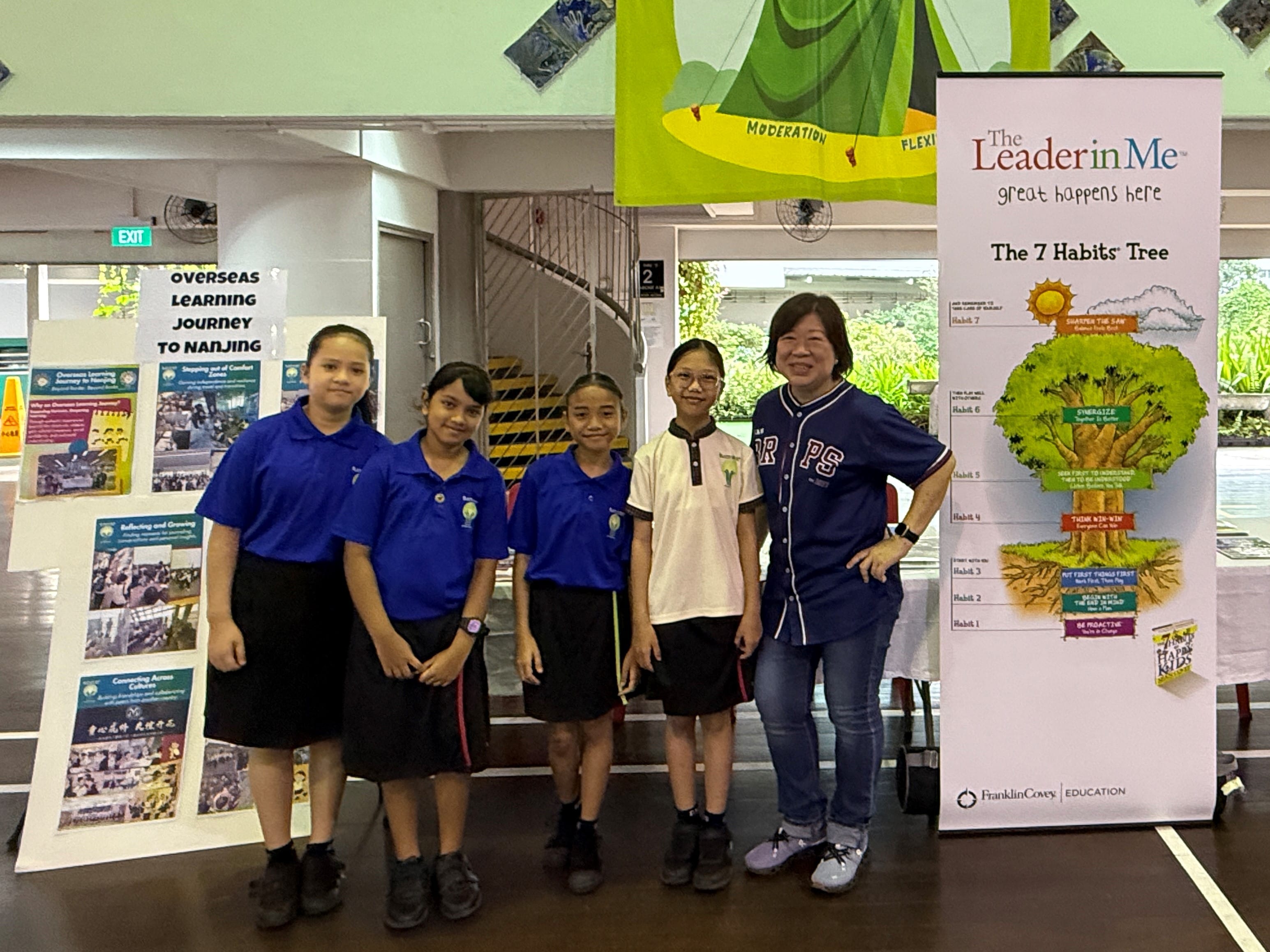Four students in school uniforms pose with a woman beside a Leader in Me 7 Habits Tree banner.