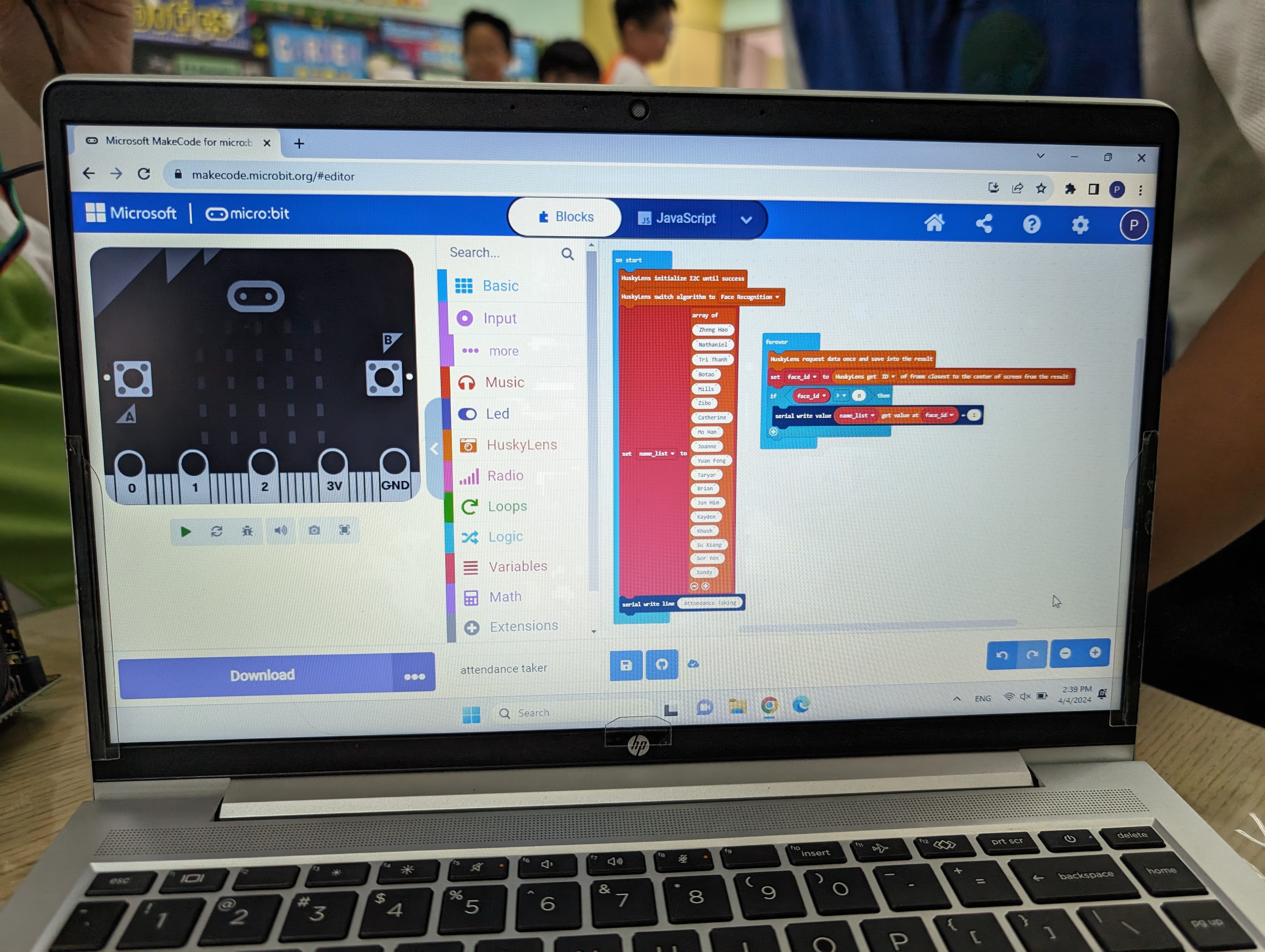Laptop showing Microsoft MakeCode for micro:bit with block code and a micro:bit simulator; blurred people in background.