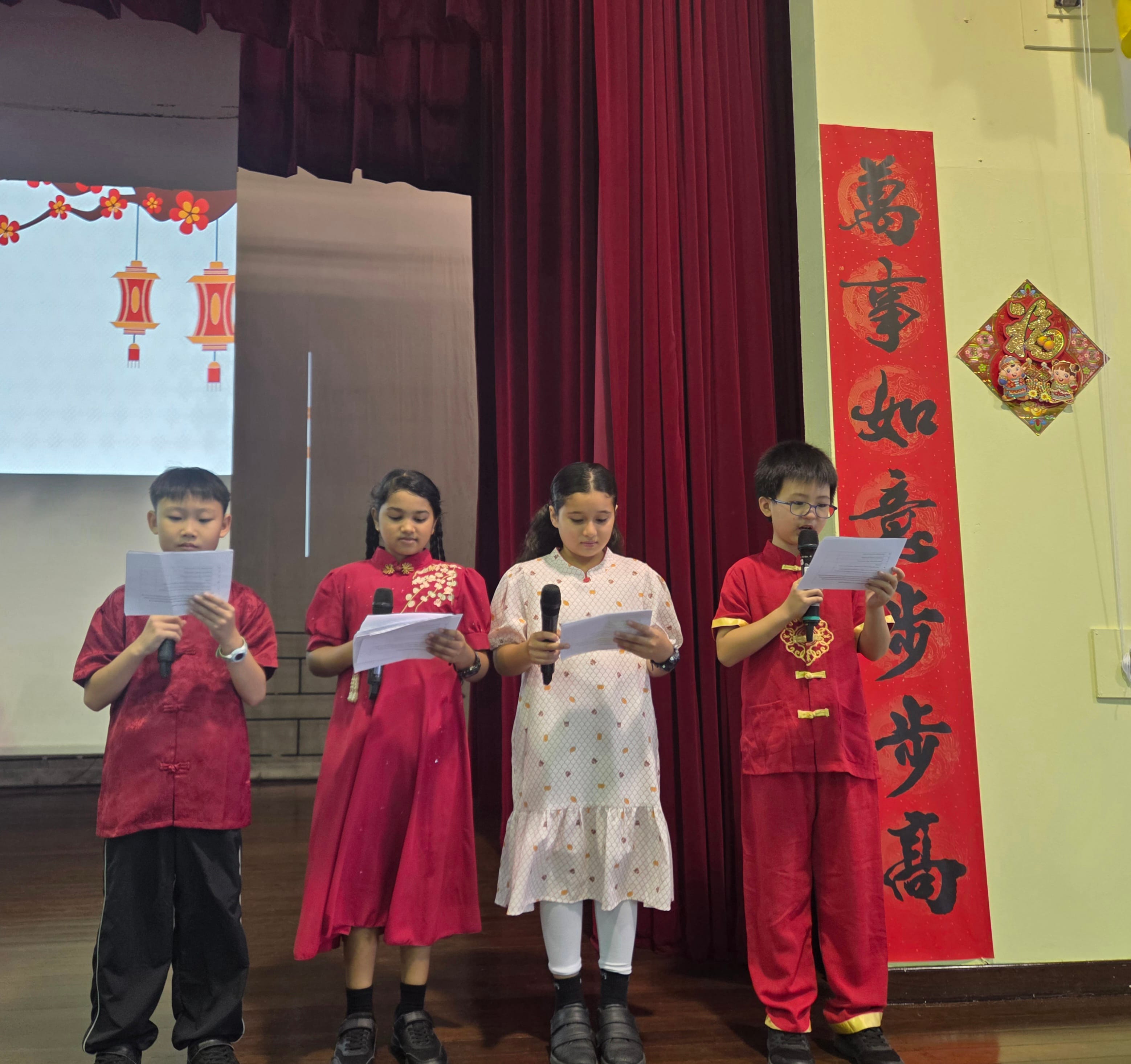 Four children hold microphones and papers on a stage decorated with Chinese New Year banners and lanterns.
