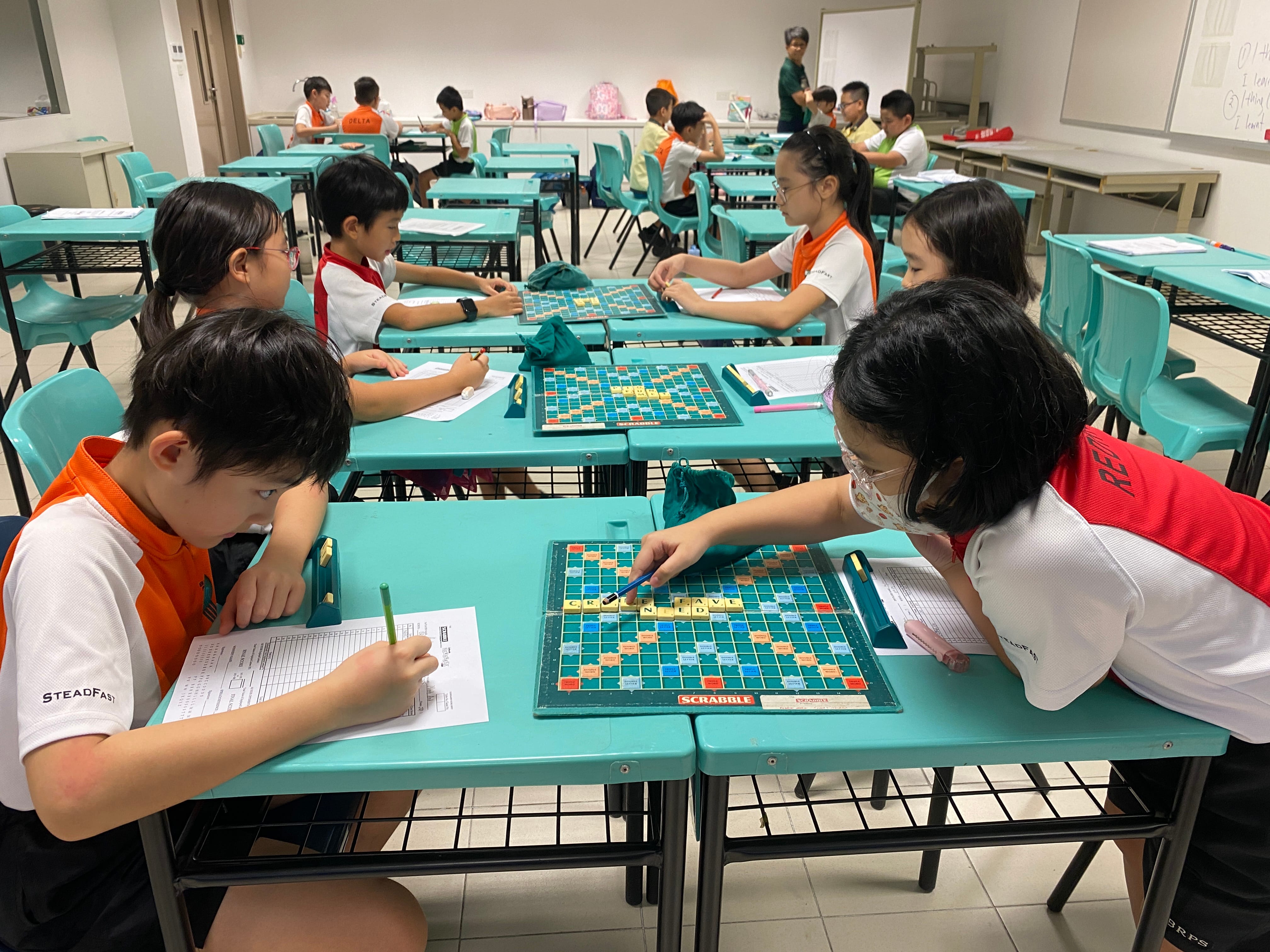 Children in school uniforms play Scrabble on teal desks in a classroom during a competition.