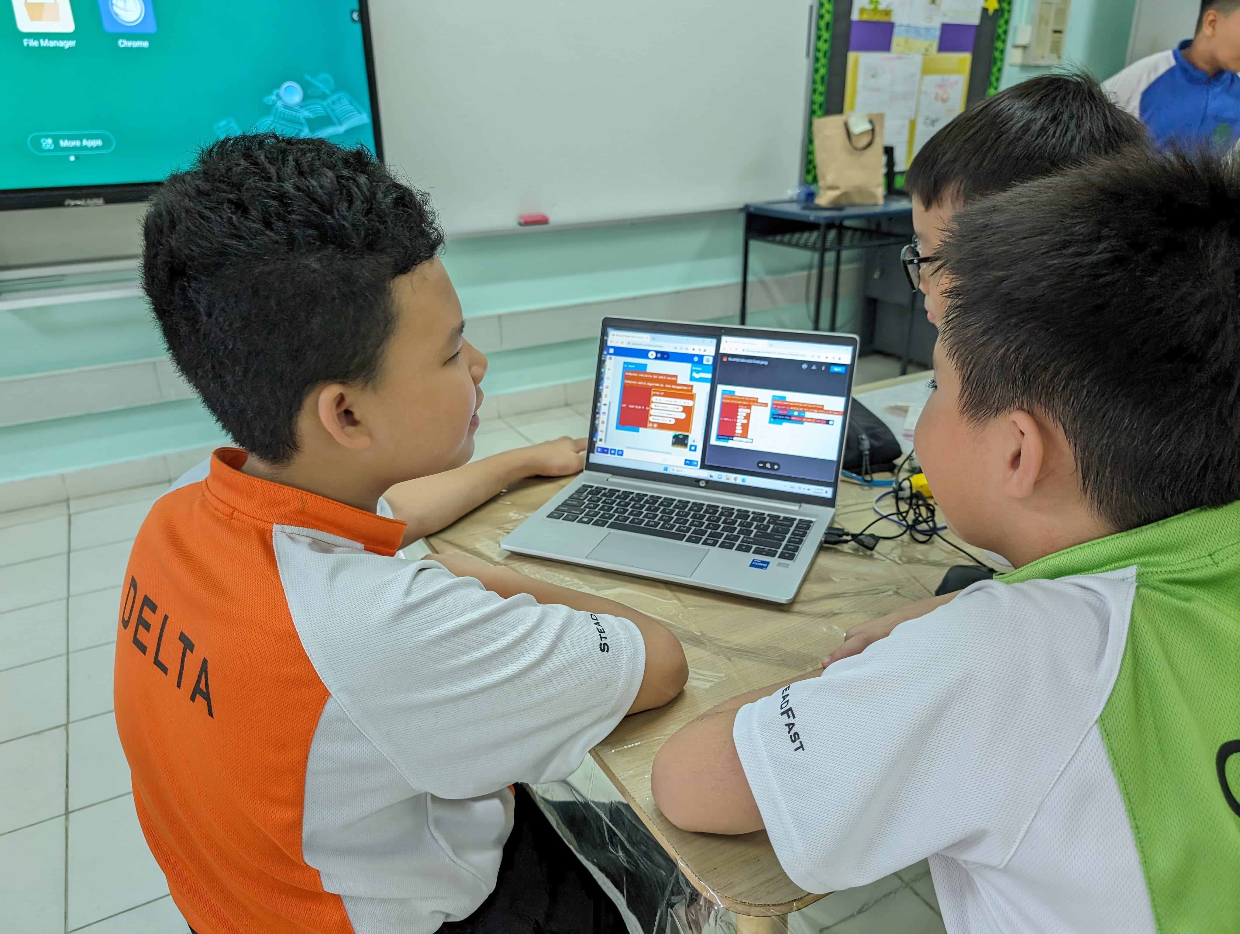 Two boys in school uniforms sit at a desk looking at a laptop showing a block-based coding program.
