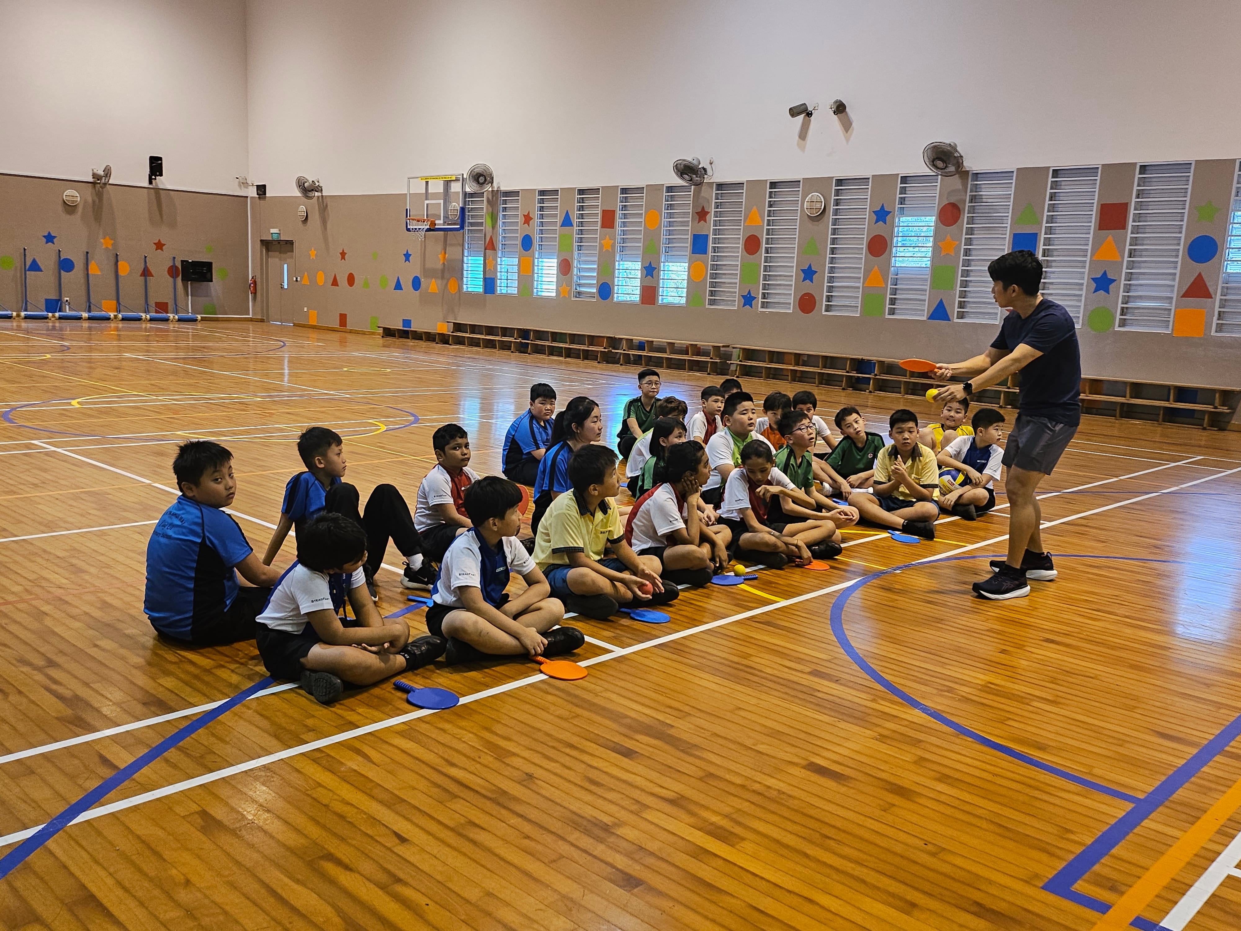 A coach holds a paddle and ball while a group of children sit on a gym floor listening during a sports lesson.