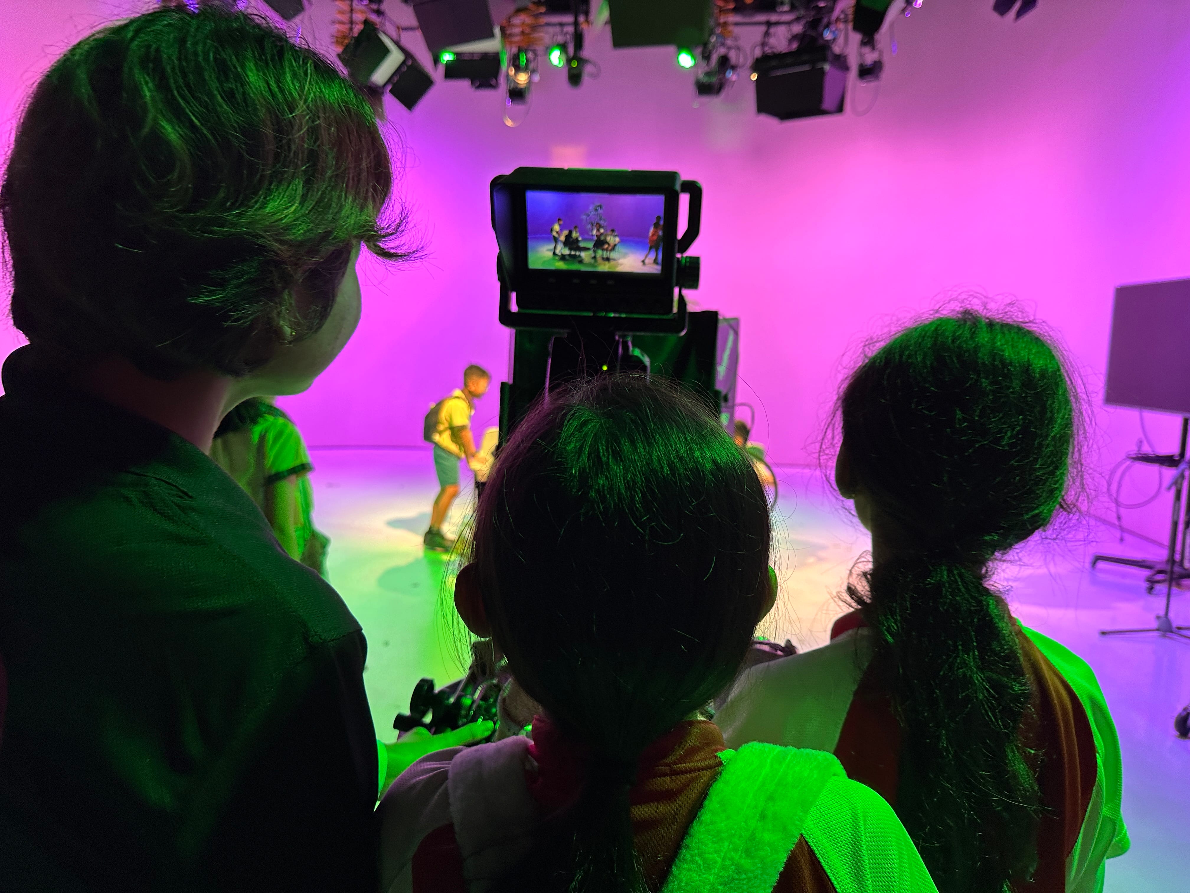 Three children from behind watch a camera monitor on a purple-lit studio set with performers in the background.