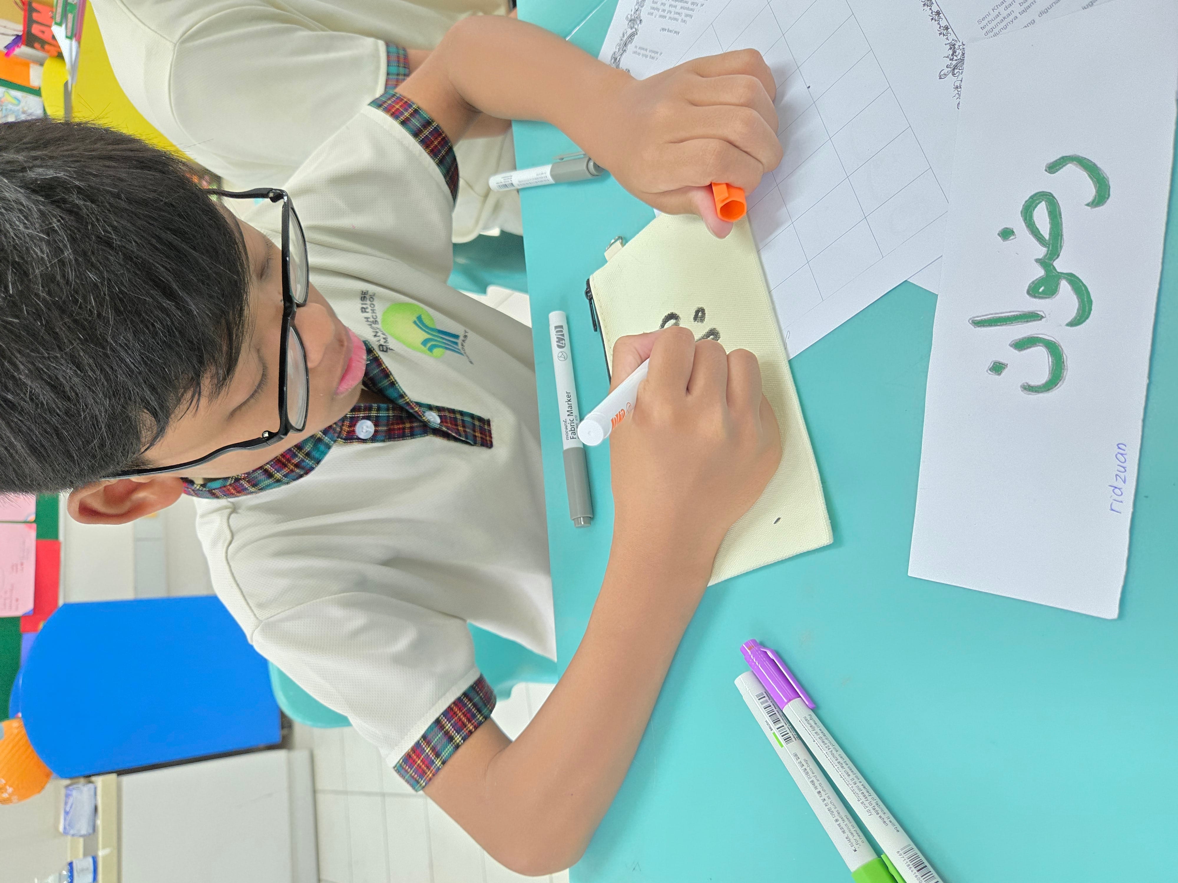 A young boy wearing glasses writes on a fabric pouch with a marker, with Arabic name card "Ridzuan" on the teal desk.