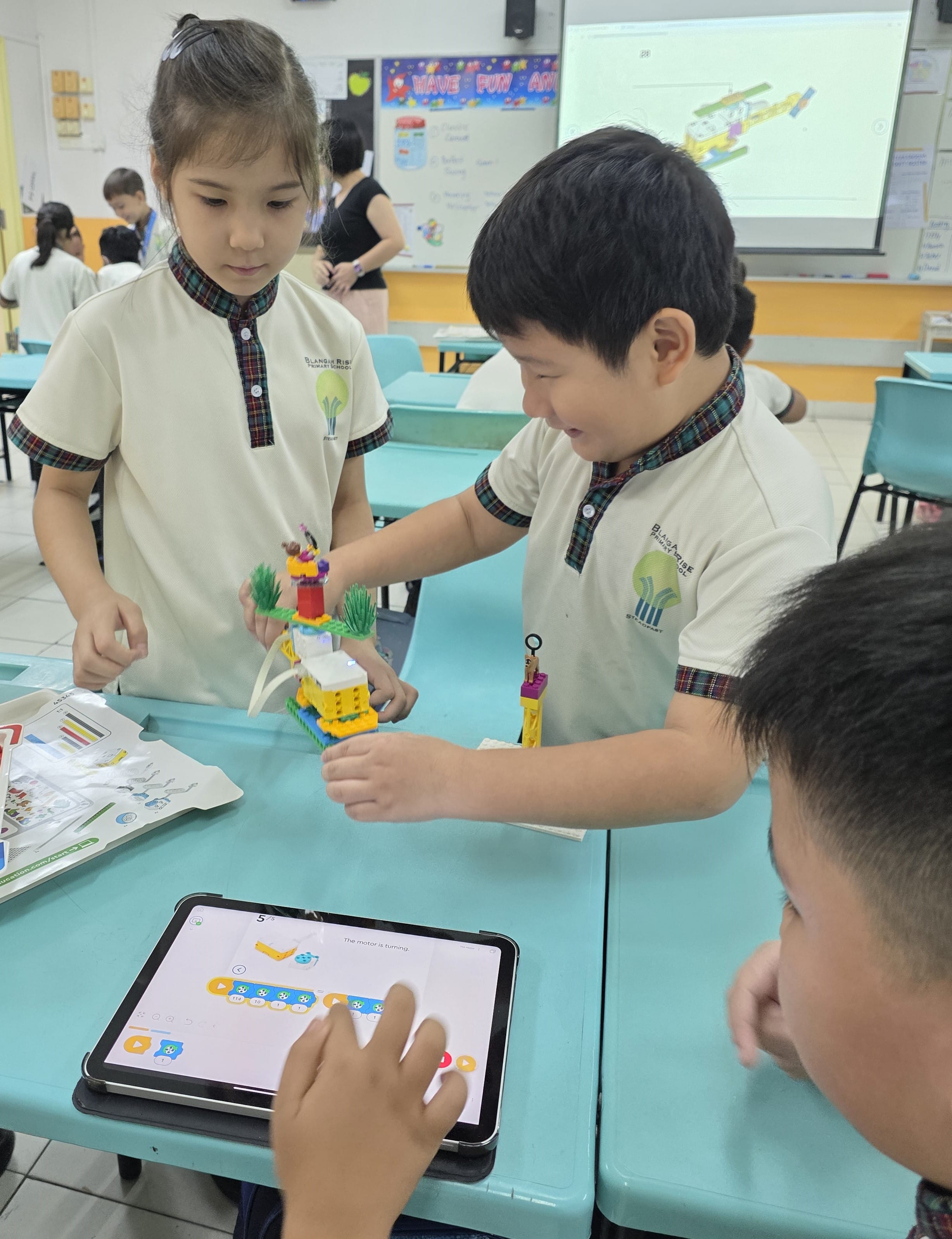 Three young students in school uniforms build a colorful LEGO model while one uses a coding tablet app in a classroom.