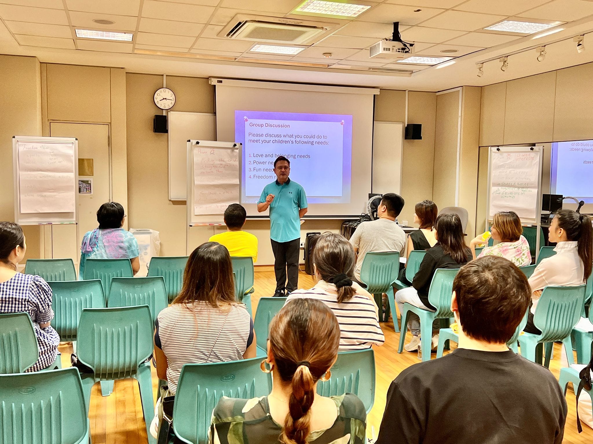 A man in a teal shirt presents a Group Discussion slide to seated attendees in a classroom with teal chairs.