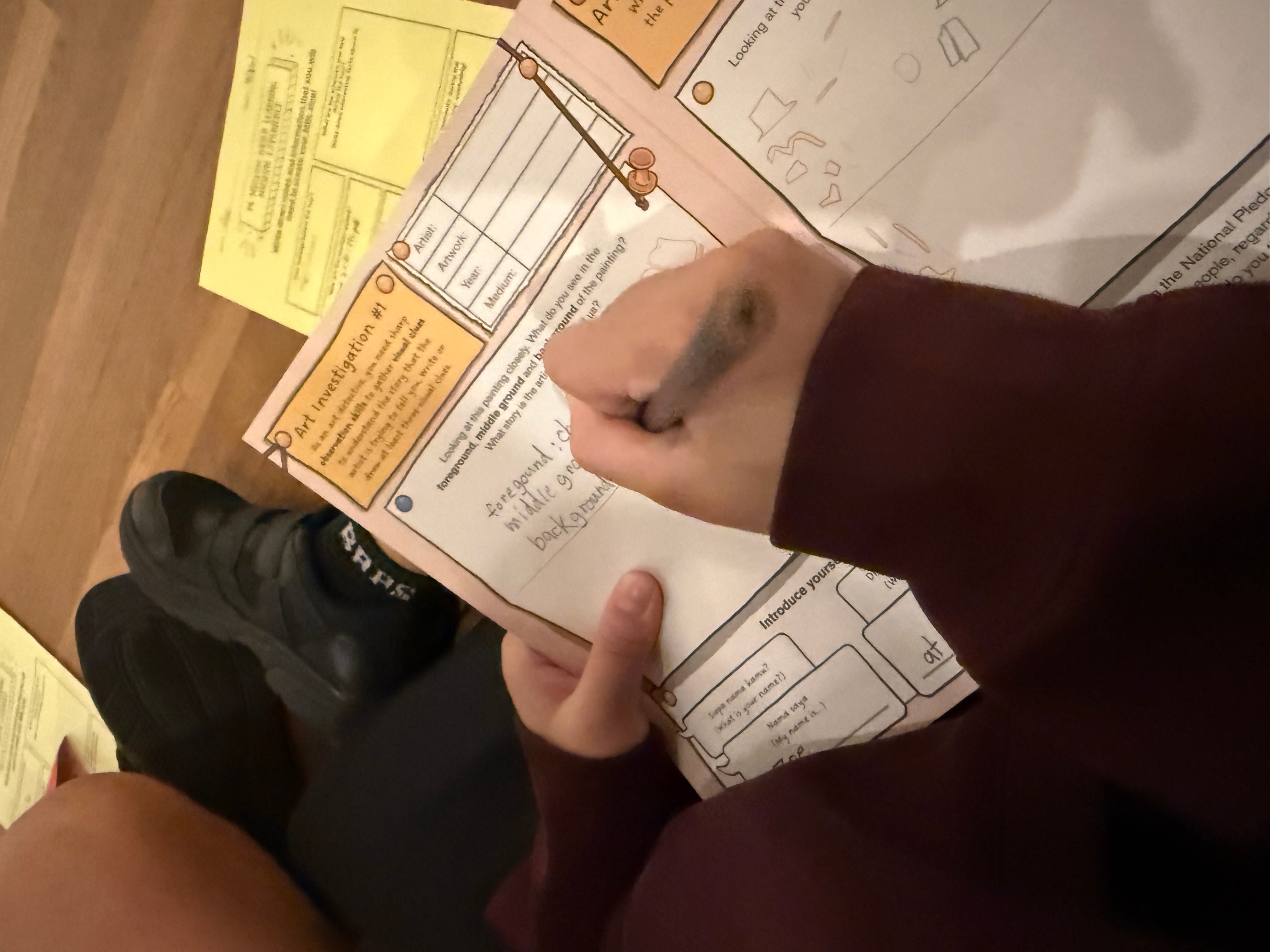 A child's hand writing on an "Art Investigation #1" worksheet with a pen while seated on a wood floor.