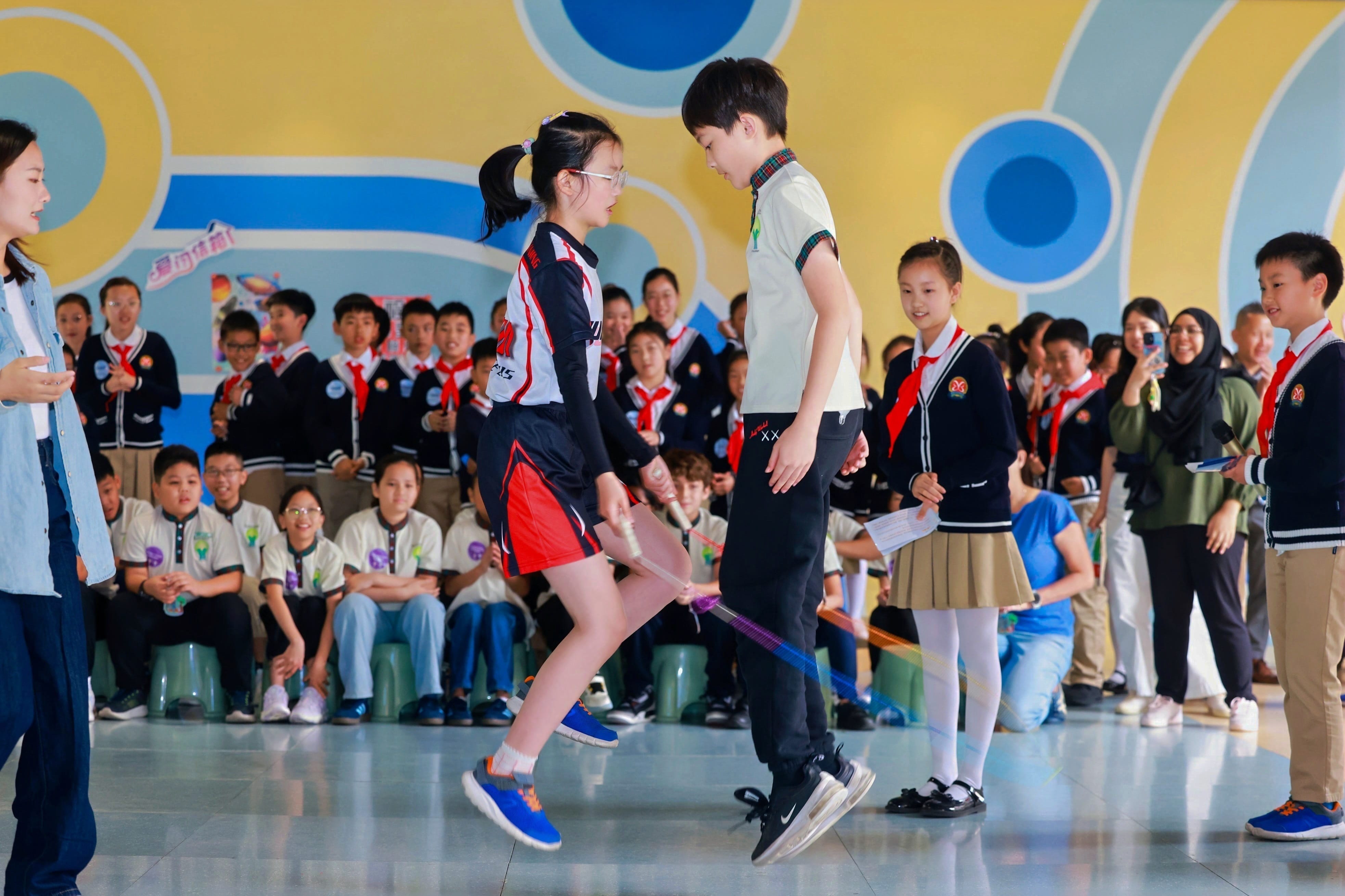 Two children jump rope in a school gym while uniformed students watch from the background.