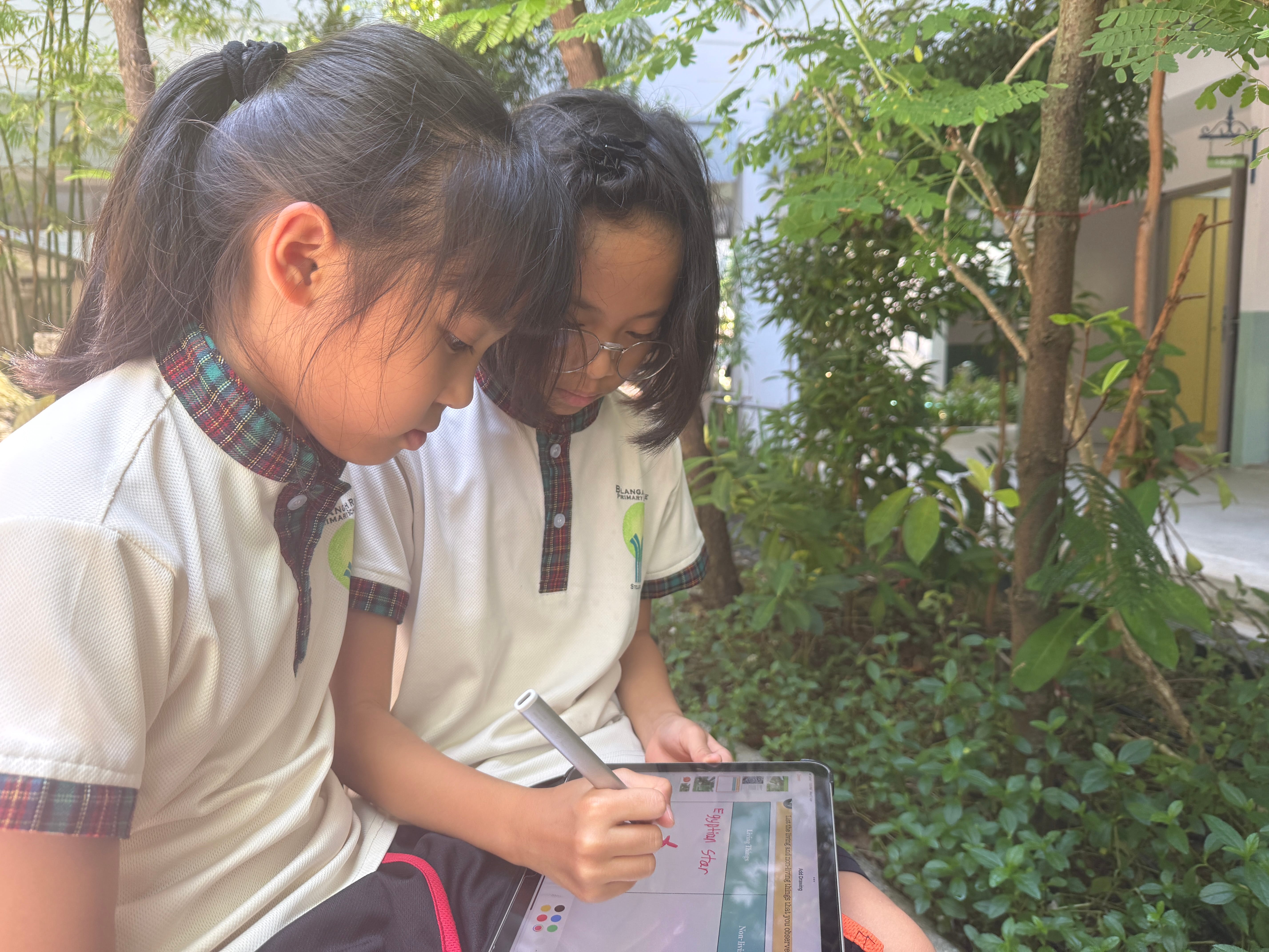 Two schoolgirls sit outdoors, looking down as one uses a stylus on a tablet amid trees and plants.