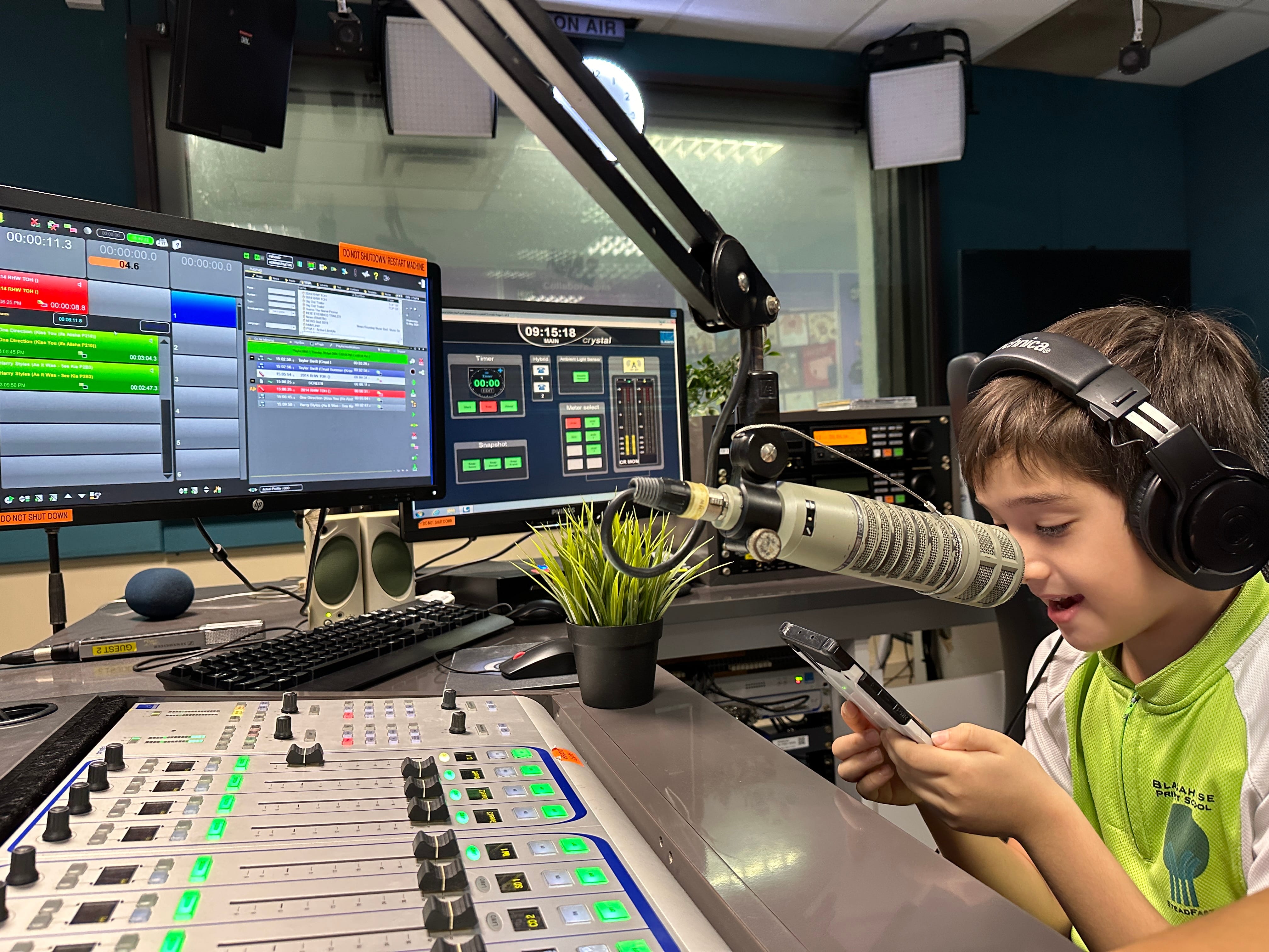 A young boy wearing headphones sits at a radio station broadcast desk with a microphone and mixing board.