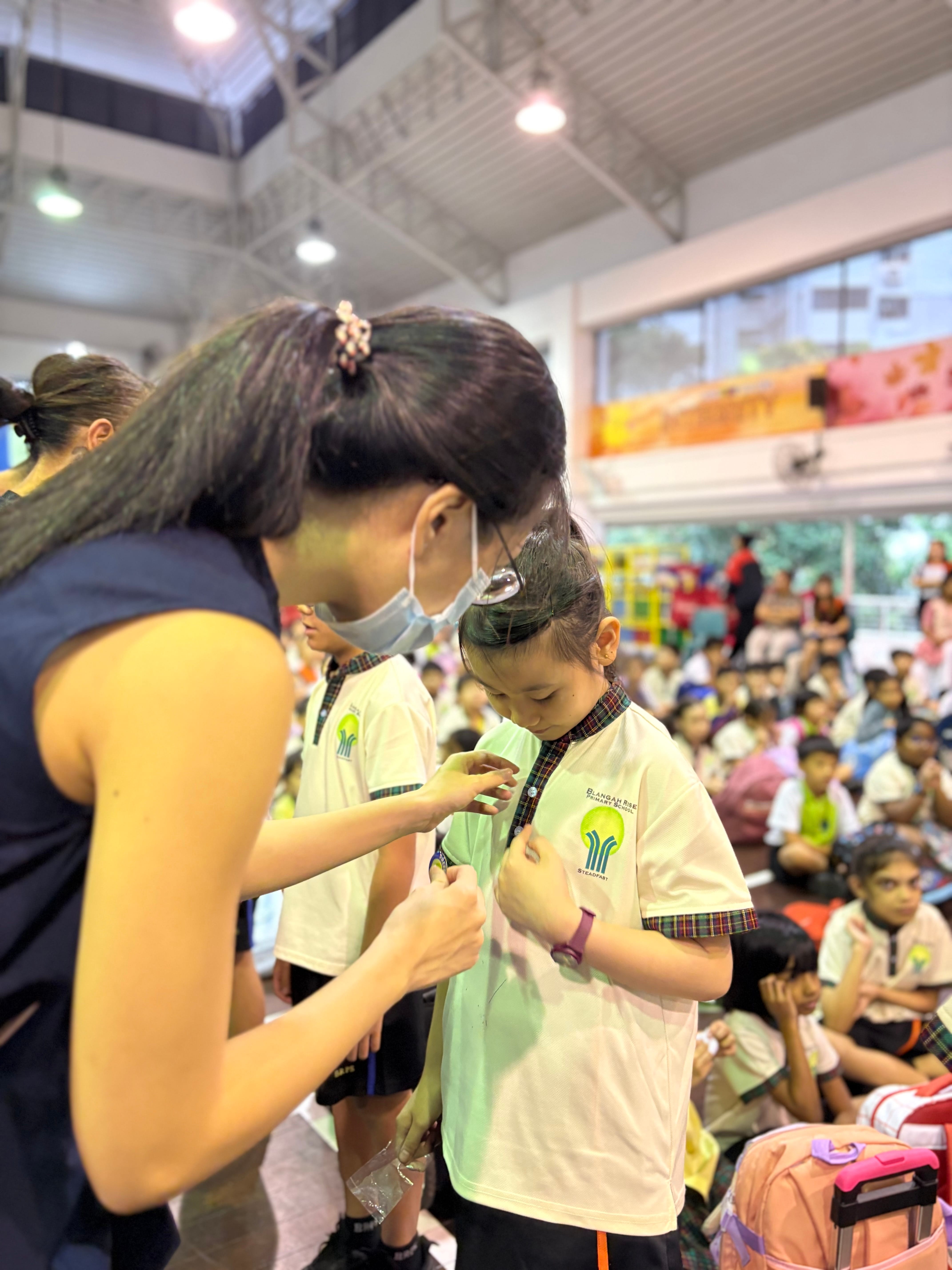 An adult pins something onto a young student's uniform in a crowded school hall with many children seated in background.