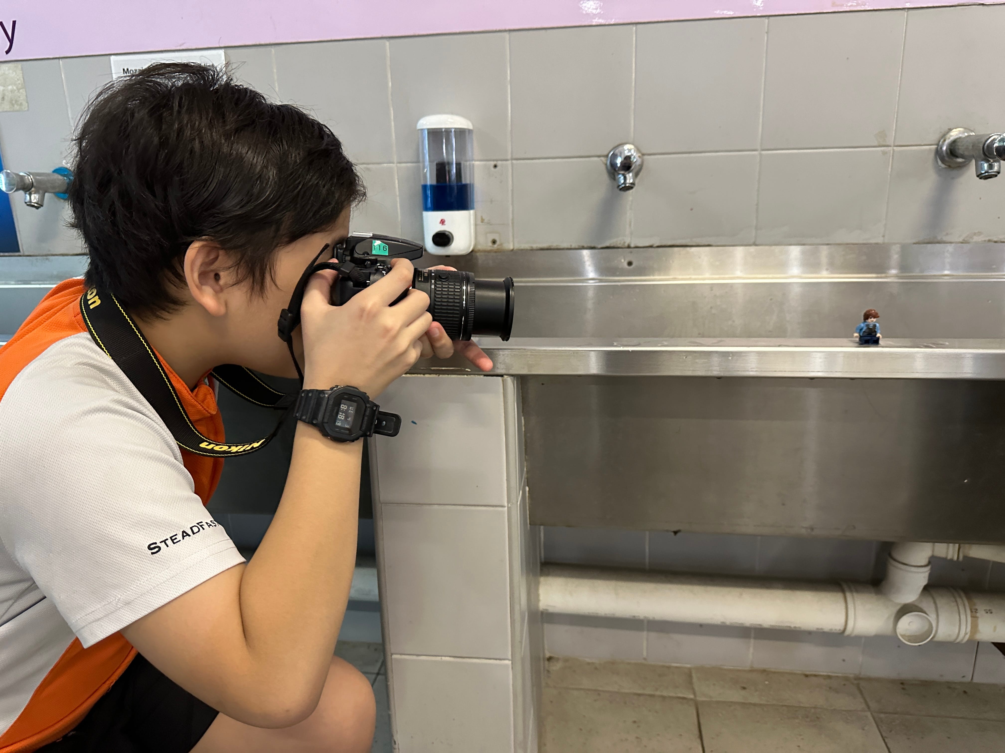 A young person crouches to photograph a small LEGO figure on a stainless steel sink with a Nikon camera.