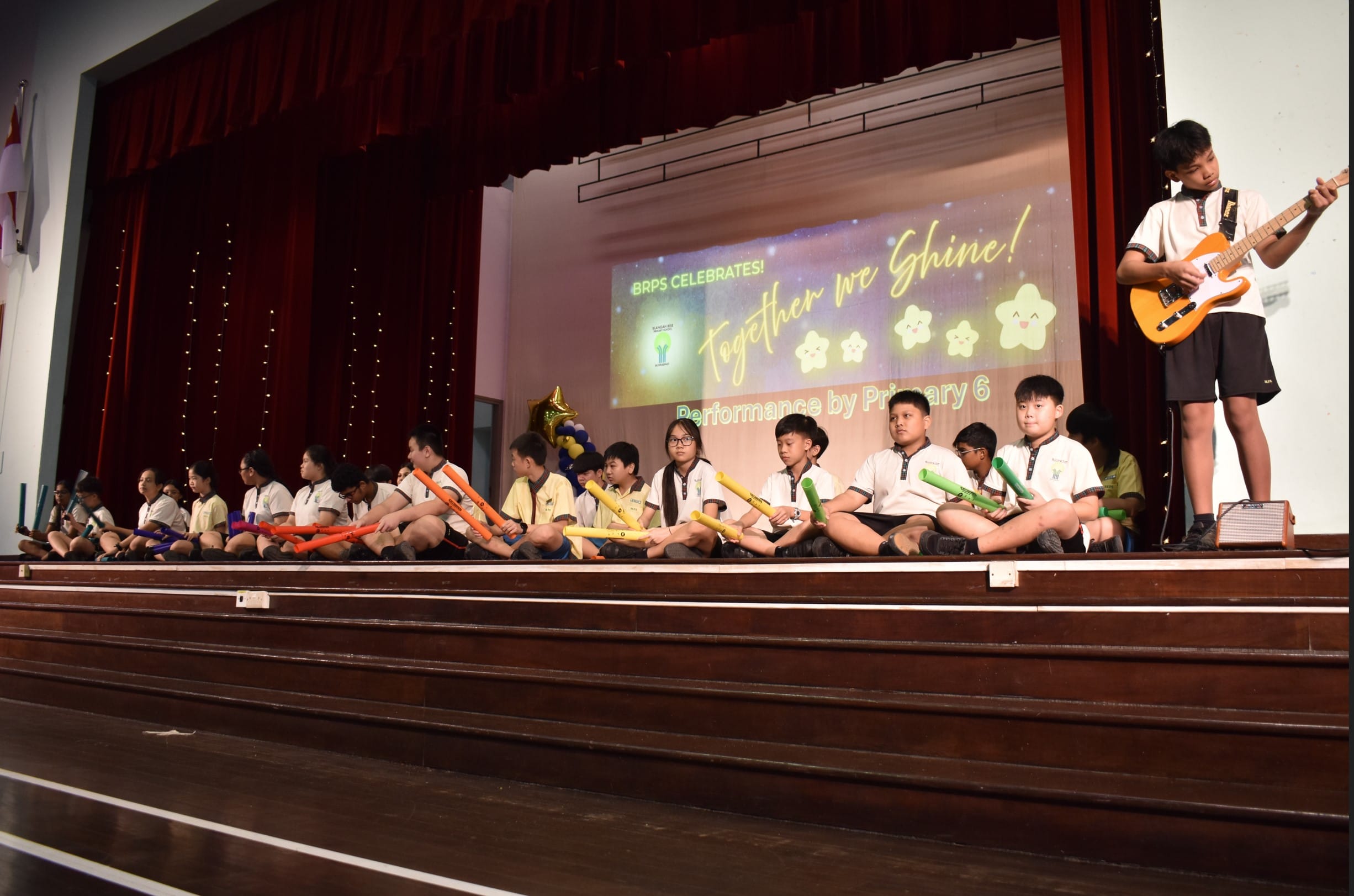 Primary 6 students sit on stage holding colorful boomwhackers while one boy stands playing a guitar at a school celebration.