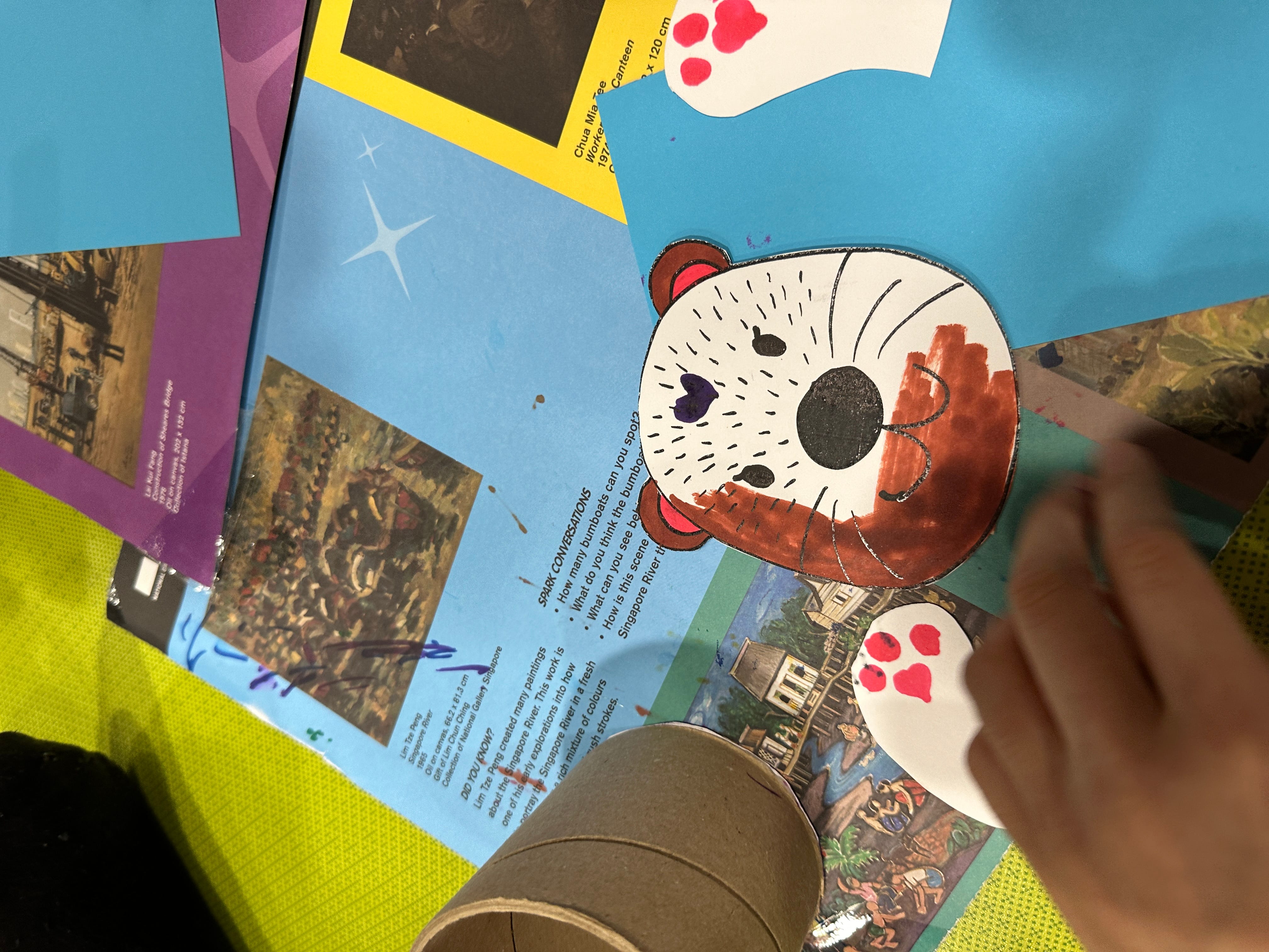 Hand paints brown and black on a paper otter face cutout on colorful paper scraps and photos.