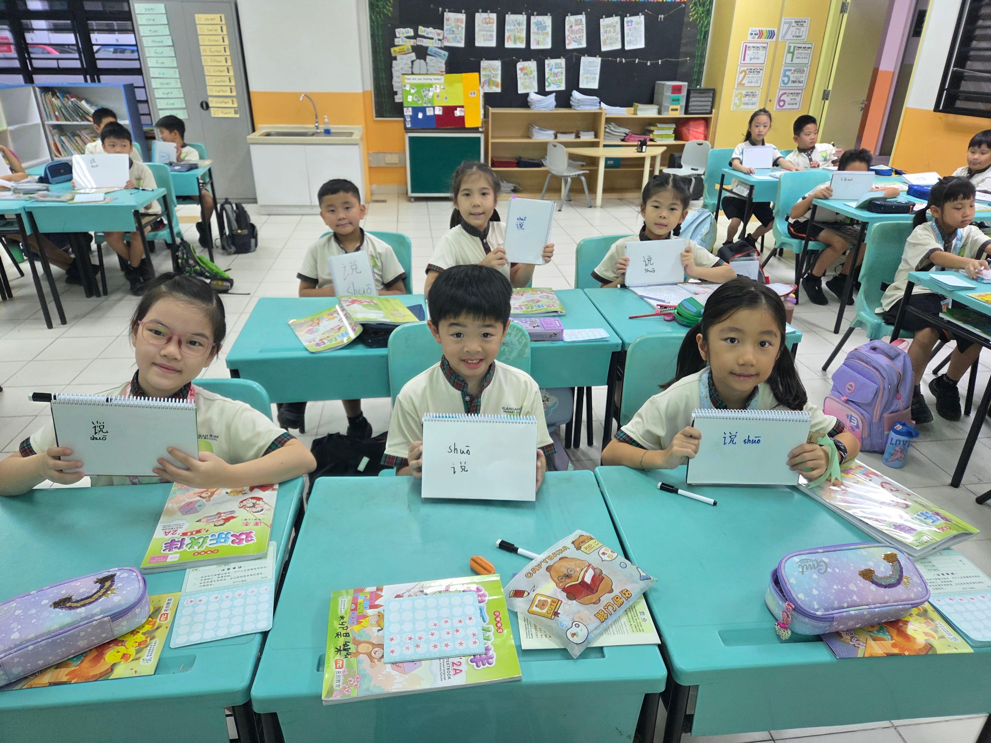 Children in a classroom hold up notebooks showing Chinese characters, seated at turquoise desks.