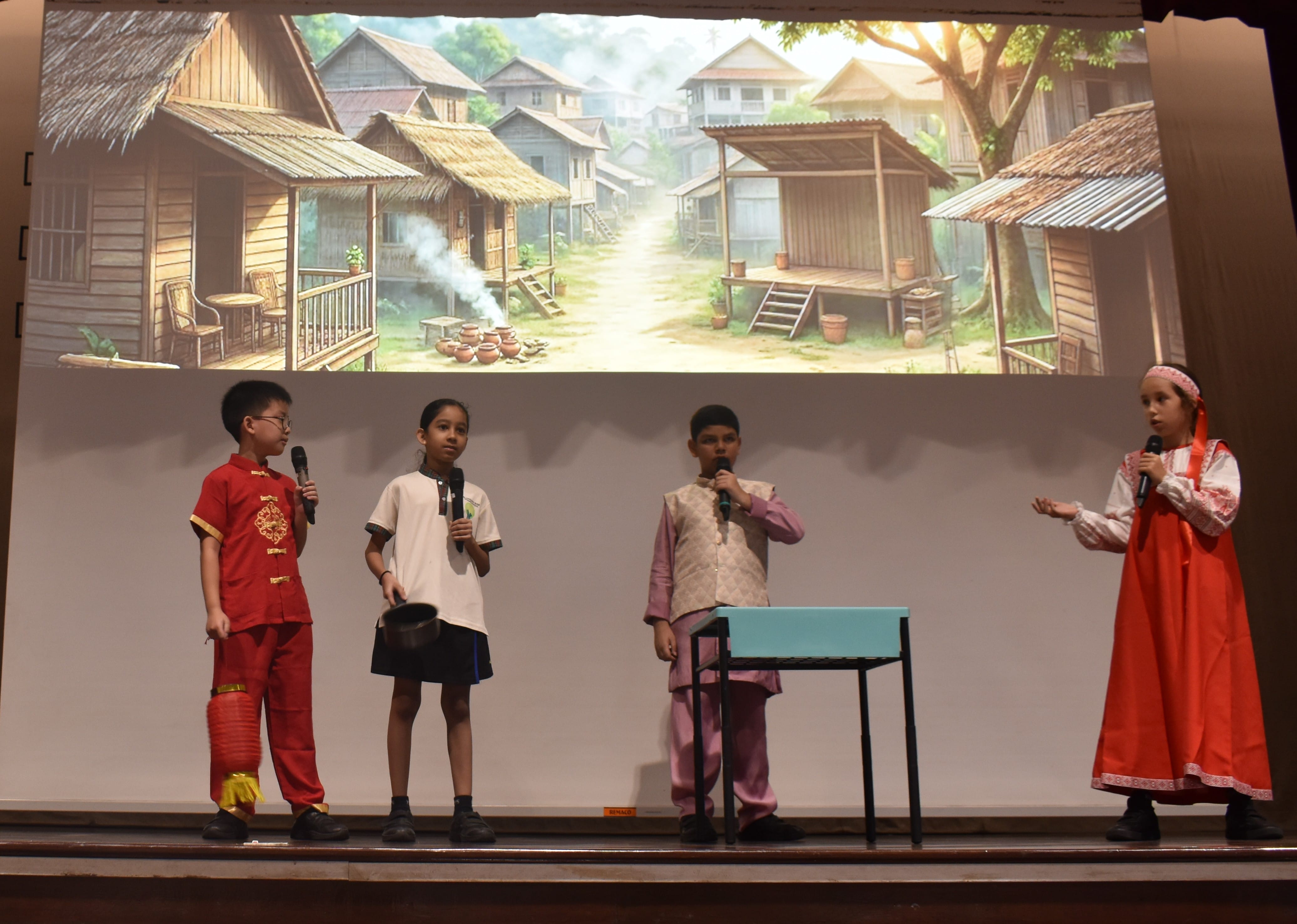 Four children in traditional cultural costumes perform on stage with microphones before a projected village backdrop.