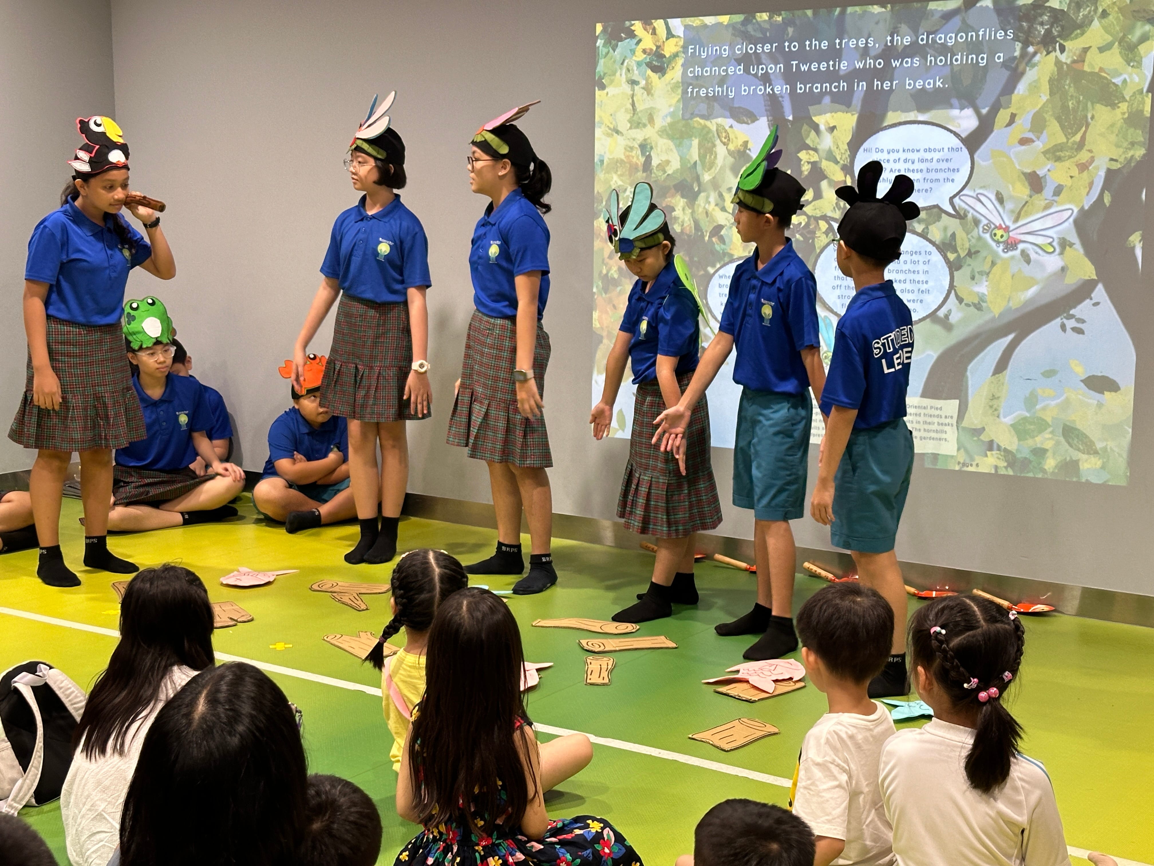 Children in costumes act out a story as classmates watch; a storybook page is projected on the wall.