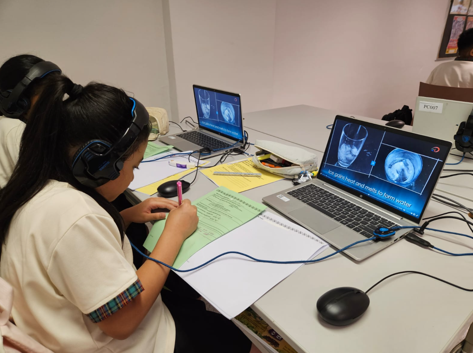 Student with headphones takes notes while watching a science lesson about ice melting on a laptop.