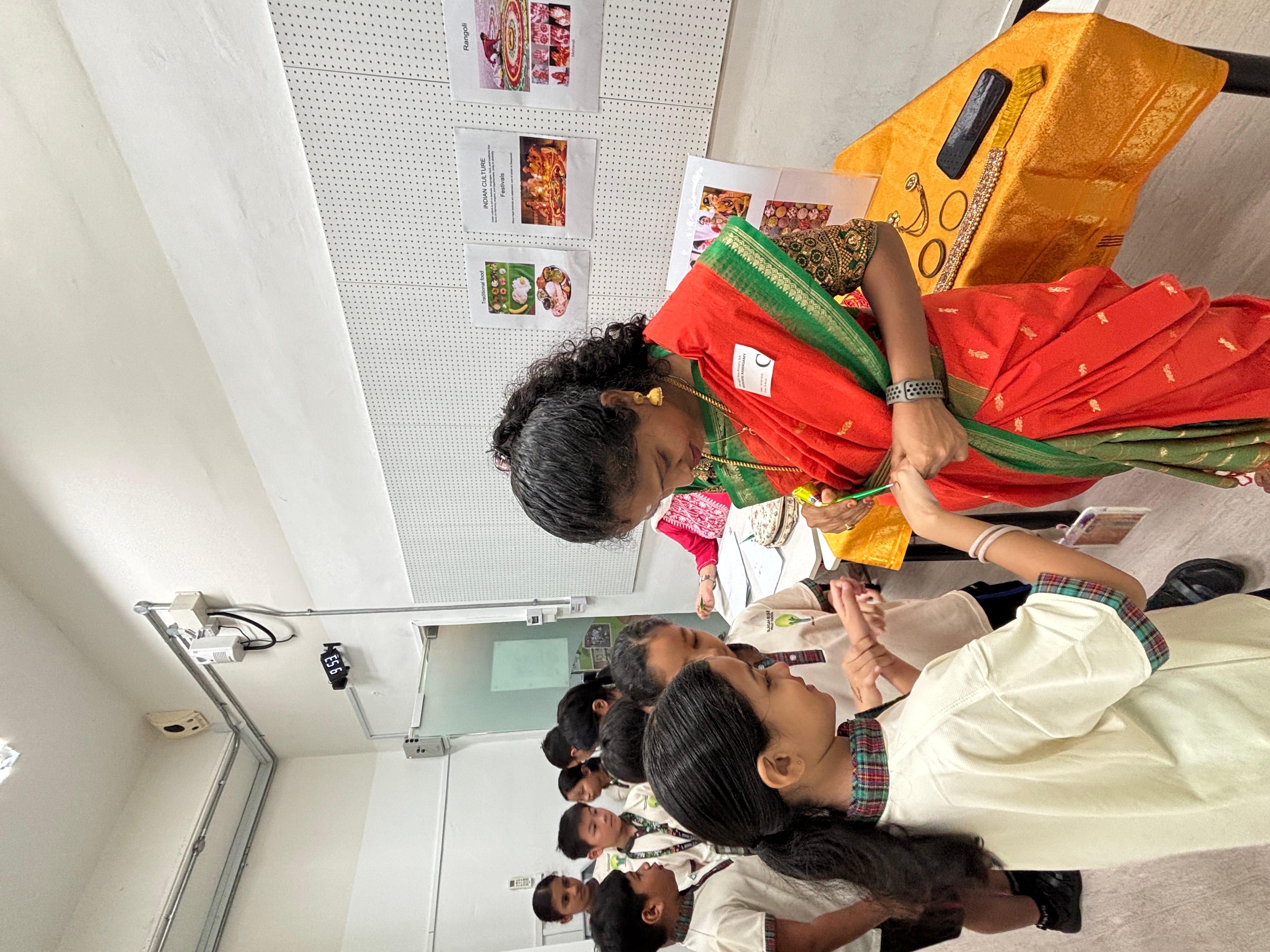 Woman in a red saree draws henna on a child's hand during an Indian culture classroom presentation.