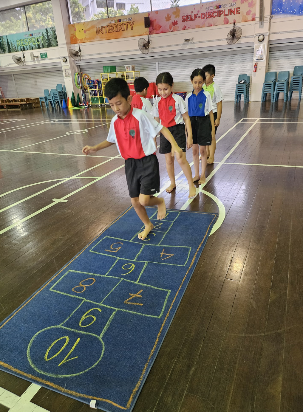 Children in school uniforms taking turns hopping on a numbered hopscotch mat in an indoor gymnasium.