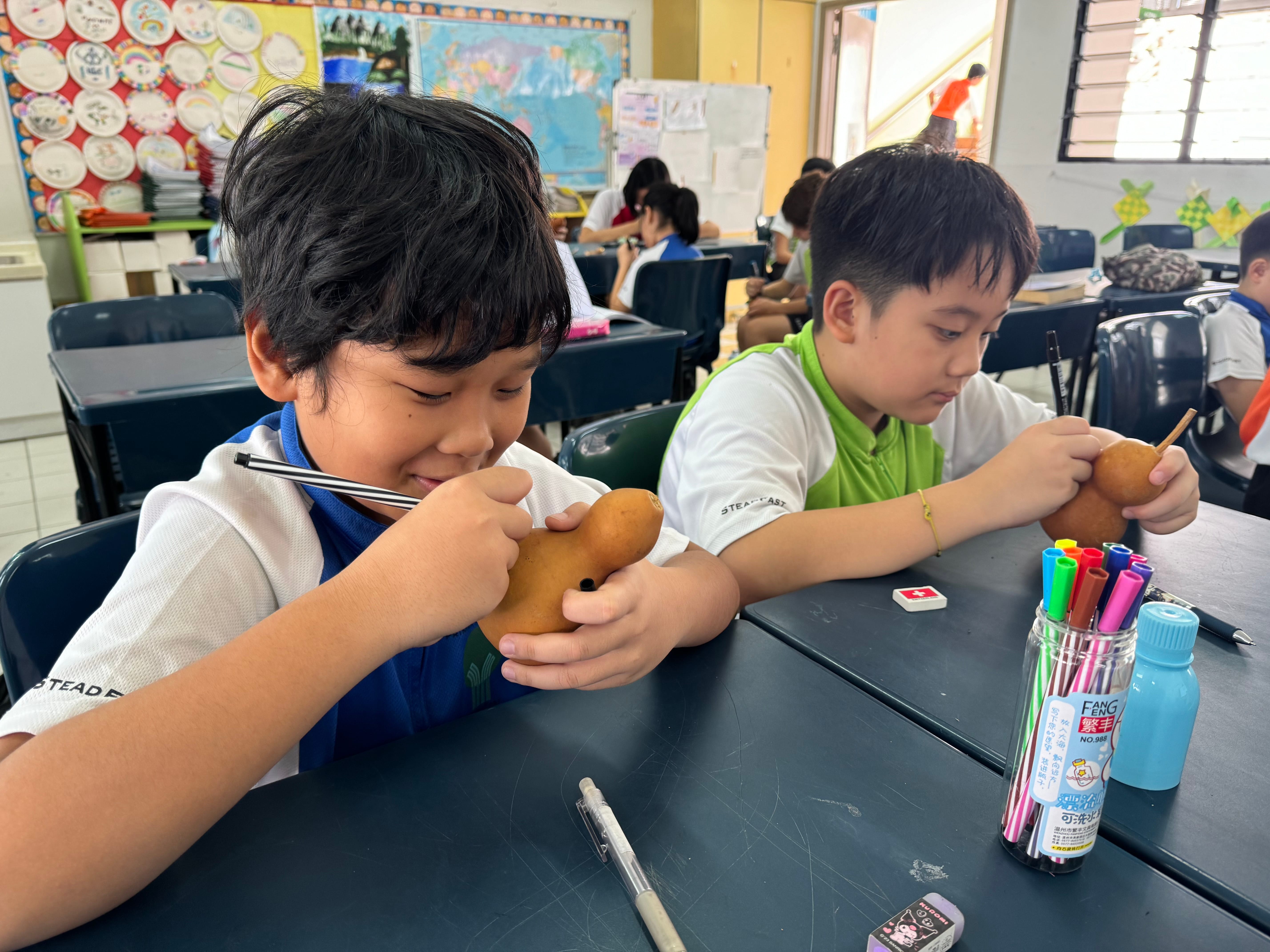 Two boys in a classroom use markers to draw on brown gourds at a desk with pens and markers.