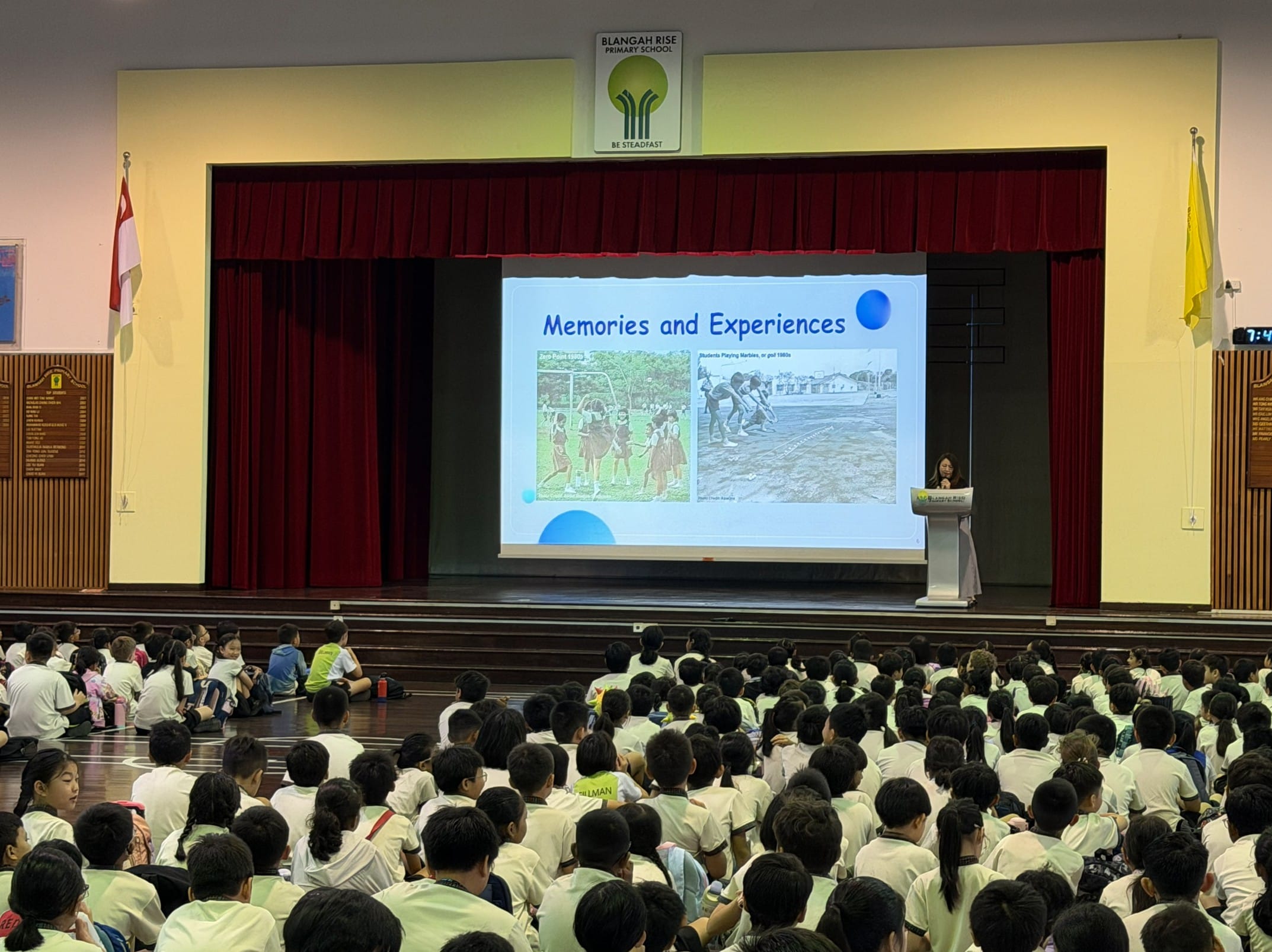 Students in white uniforms sit on a hall floor watching a "Memories and Experiences" slideshow at Blangah Rise Primary School.