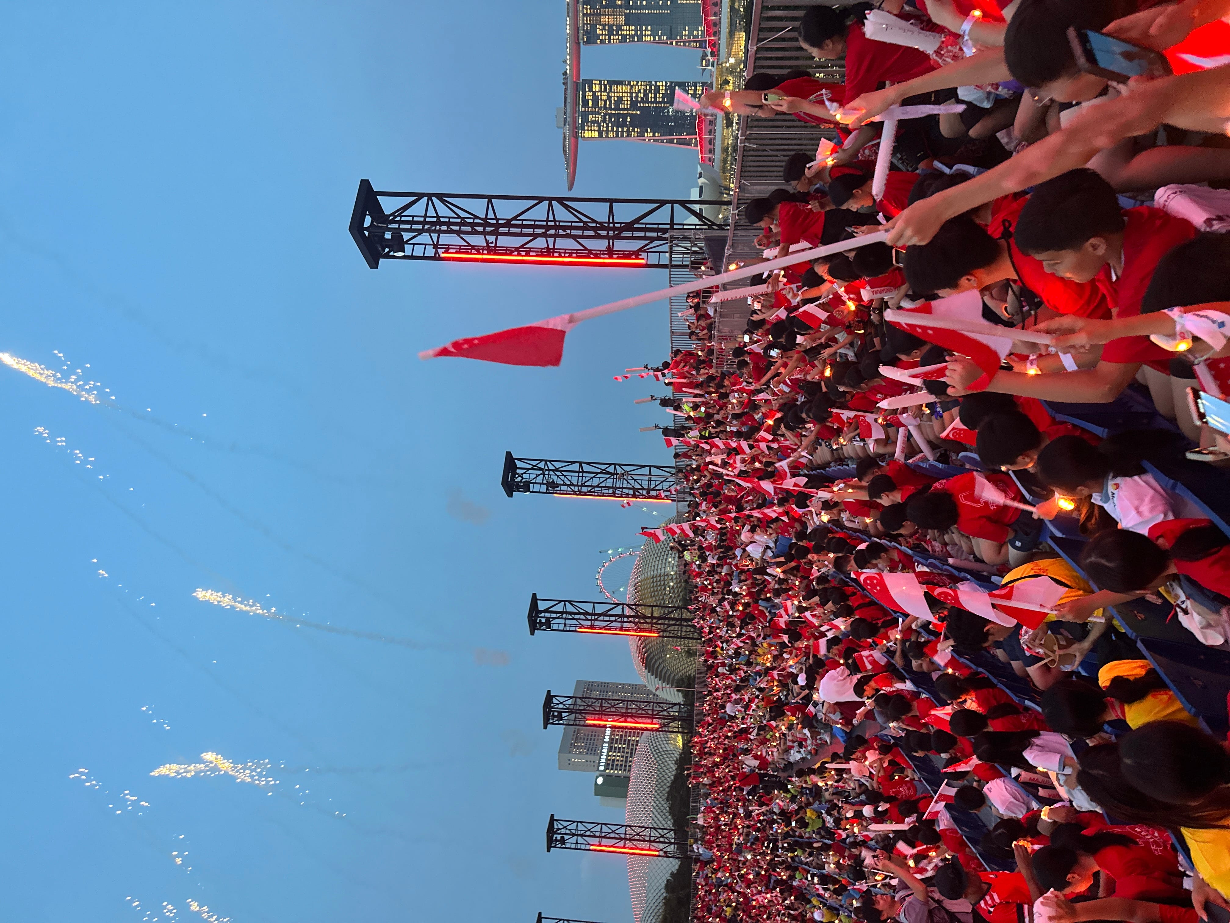 Large crowd in red shirts waving Singapore flags at an outdoor event with fireworks launching into the dusk sky.
