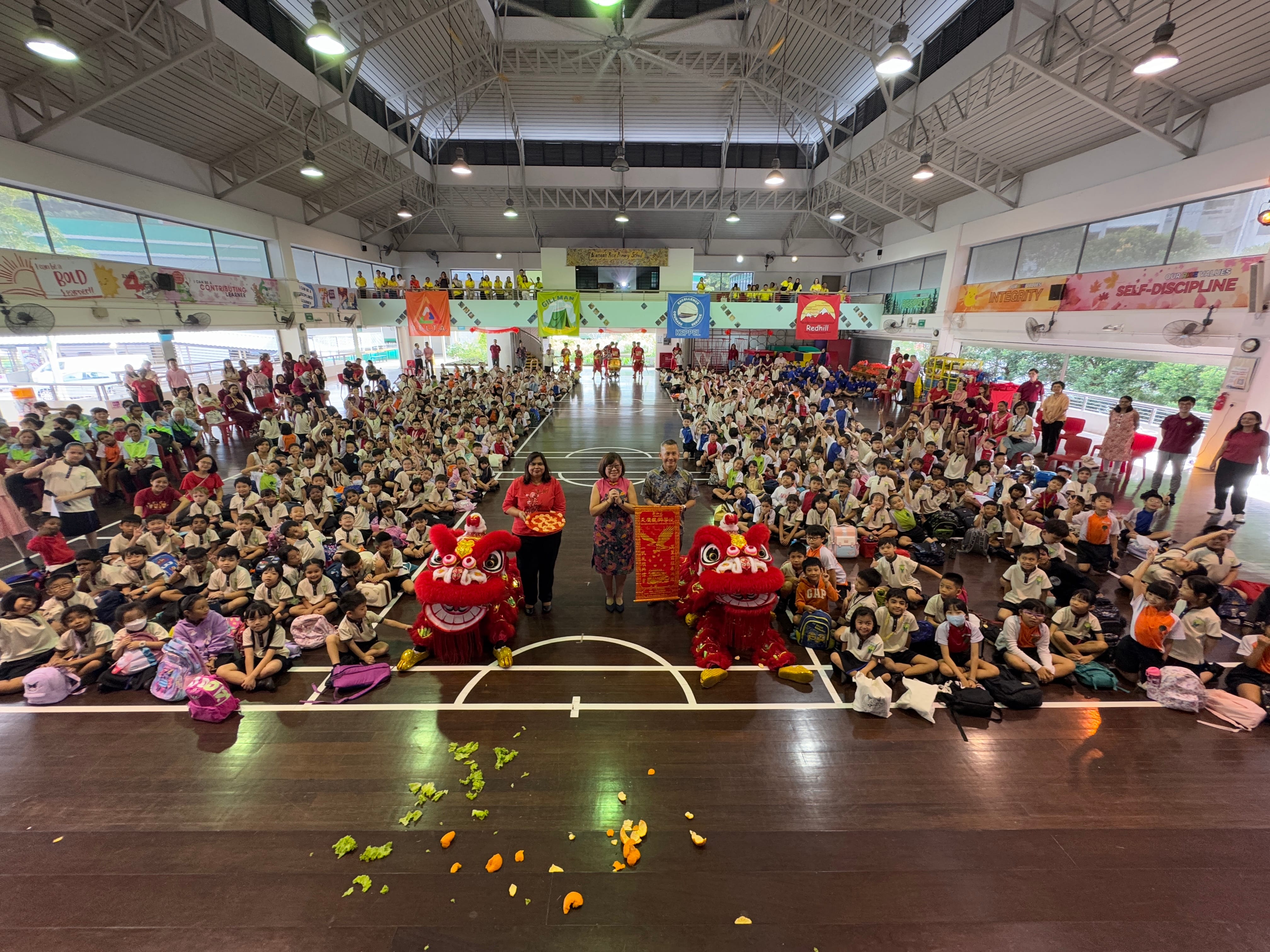 Large school hall filled with students watching a Chinese lion dance celebration with two red lion costumes and adults posing.