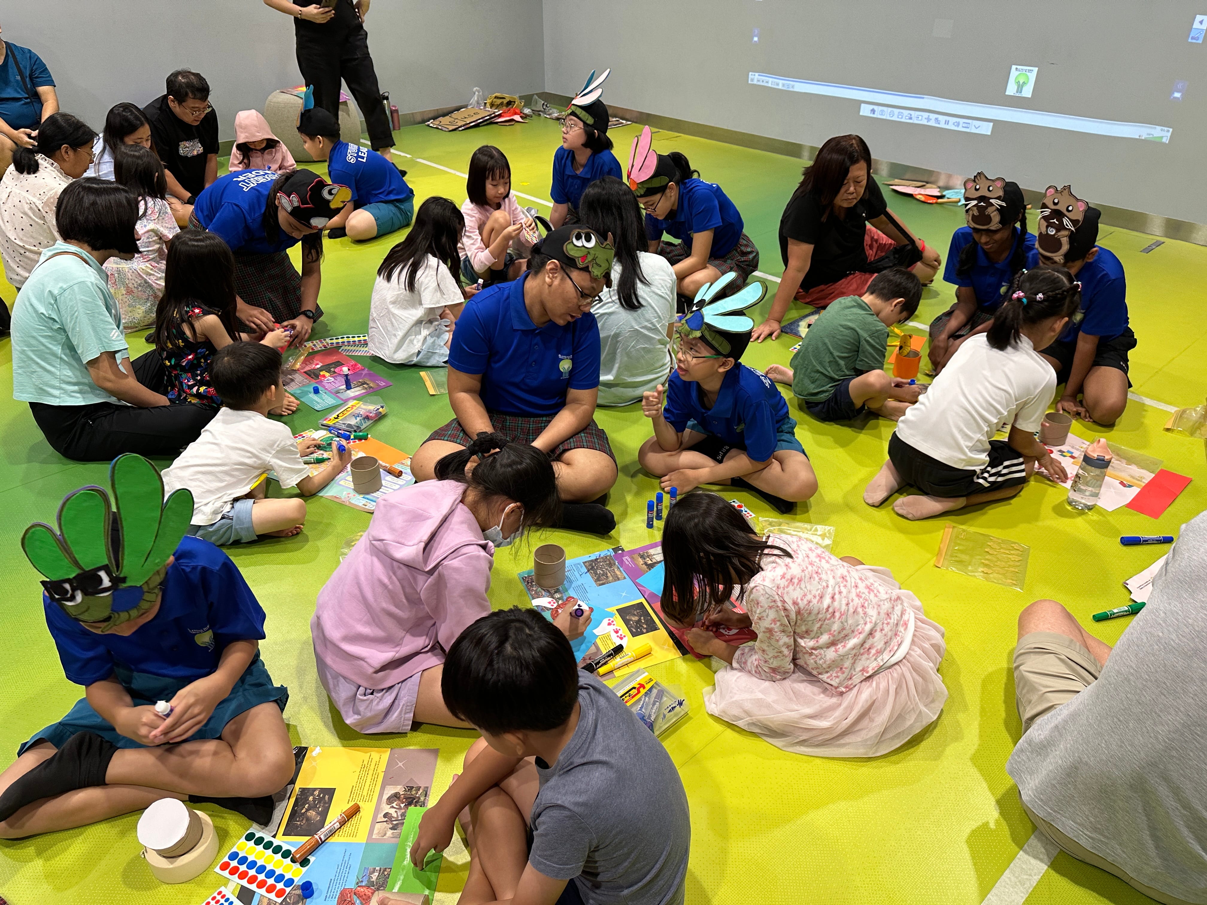 Children and adults sit on a green floor doing crafts, some wearing animal and insect headbands.