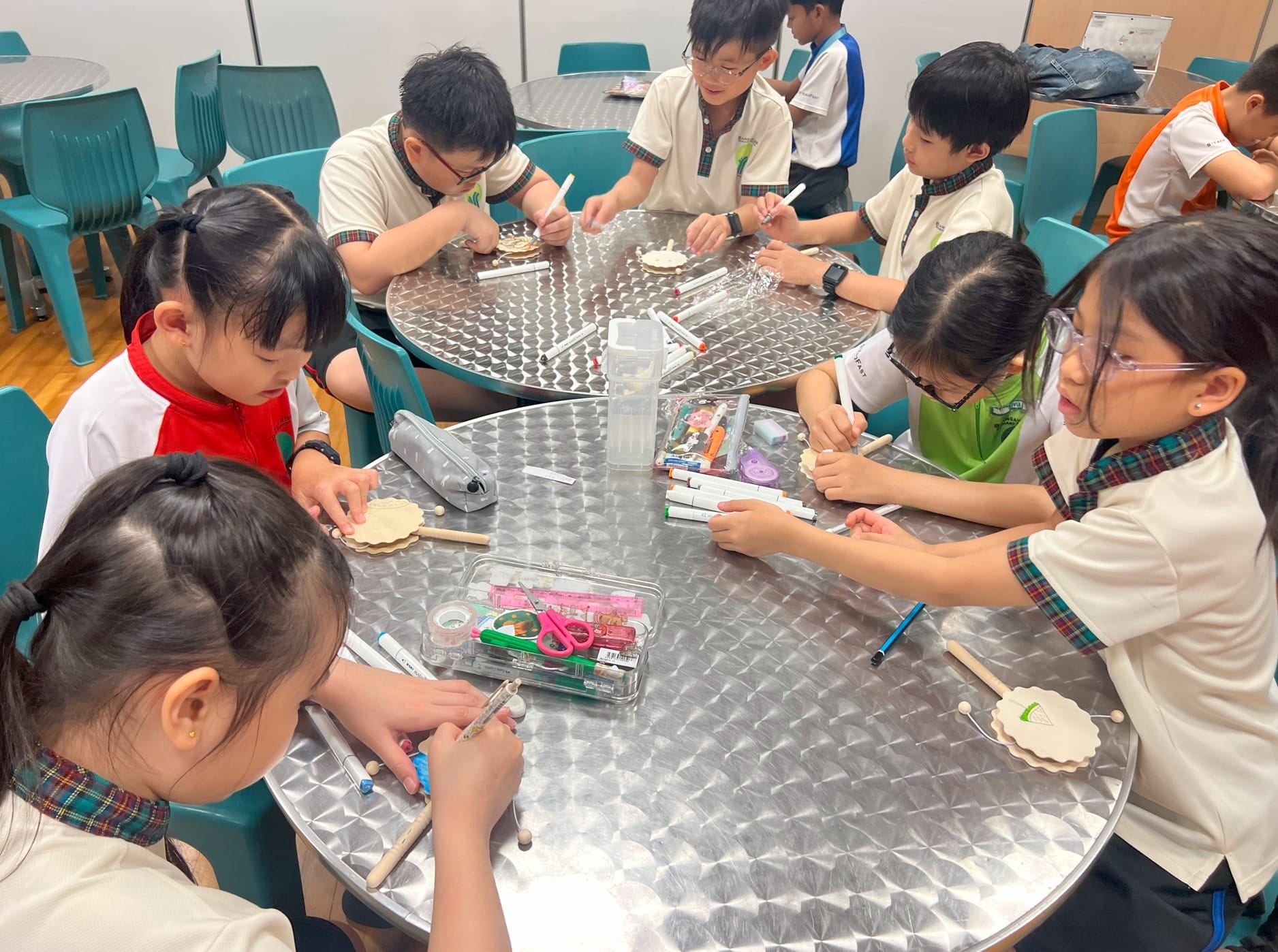 Several children in school uniforms sit around a round table doing a craft activity with markers and wooden pieces.