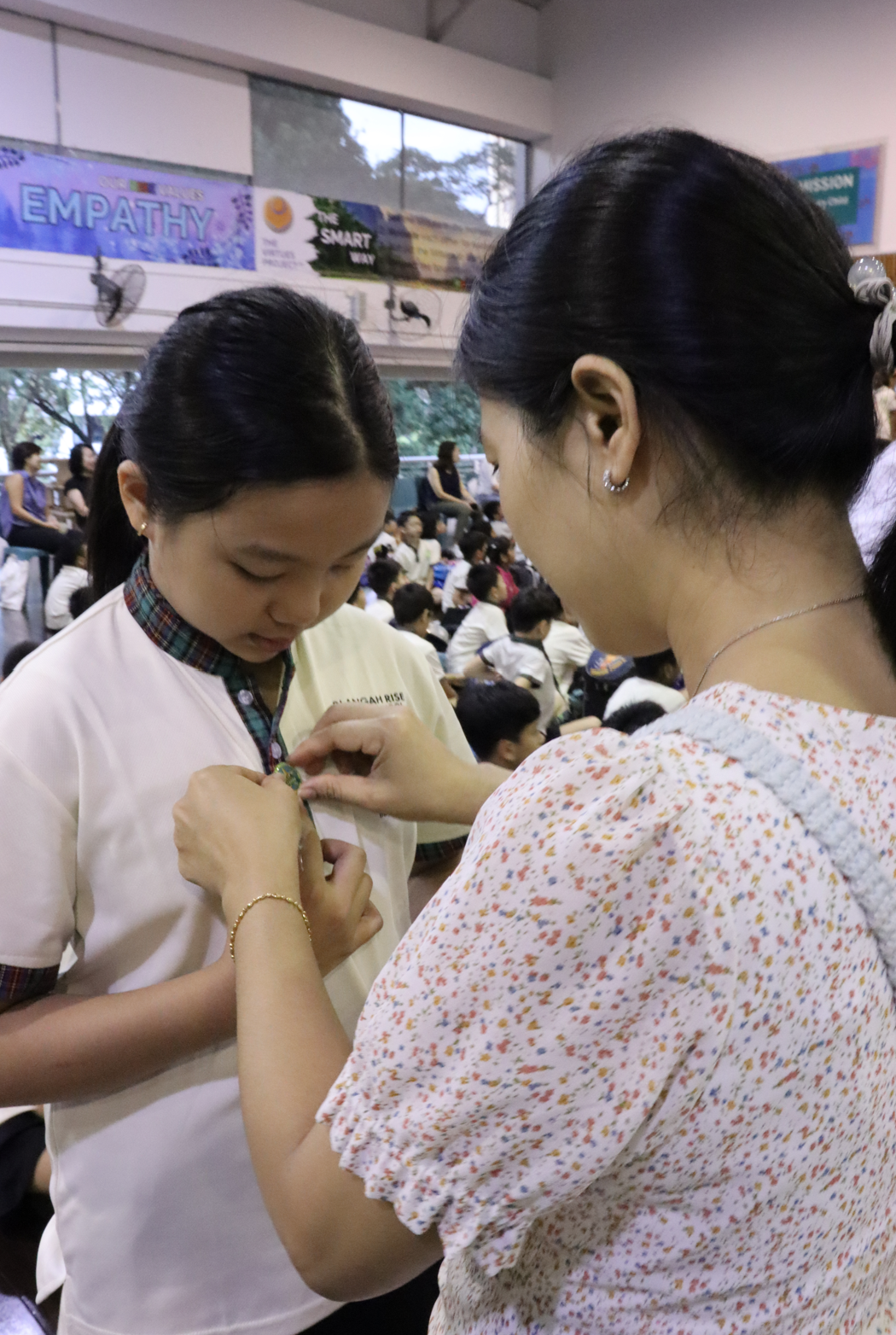 Two young women pin a badge onto a school uniform in a crowded hall with an Empathy banner on the wall.