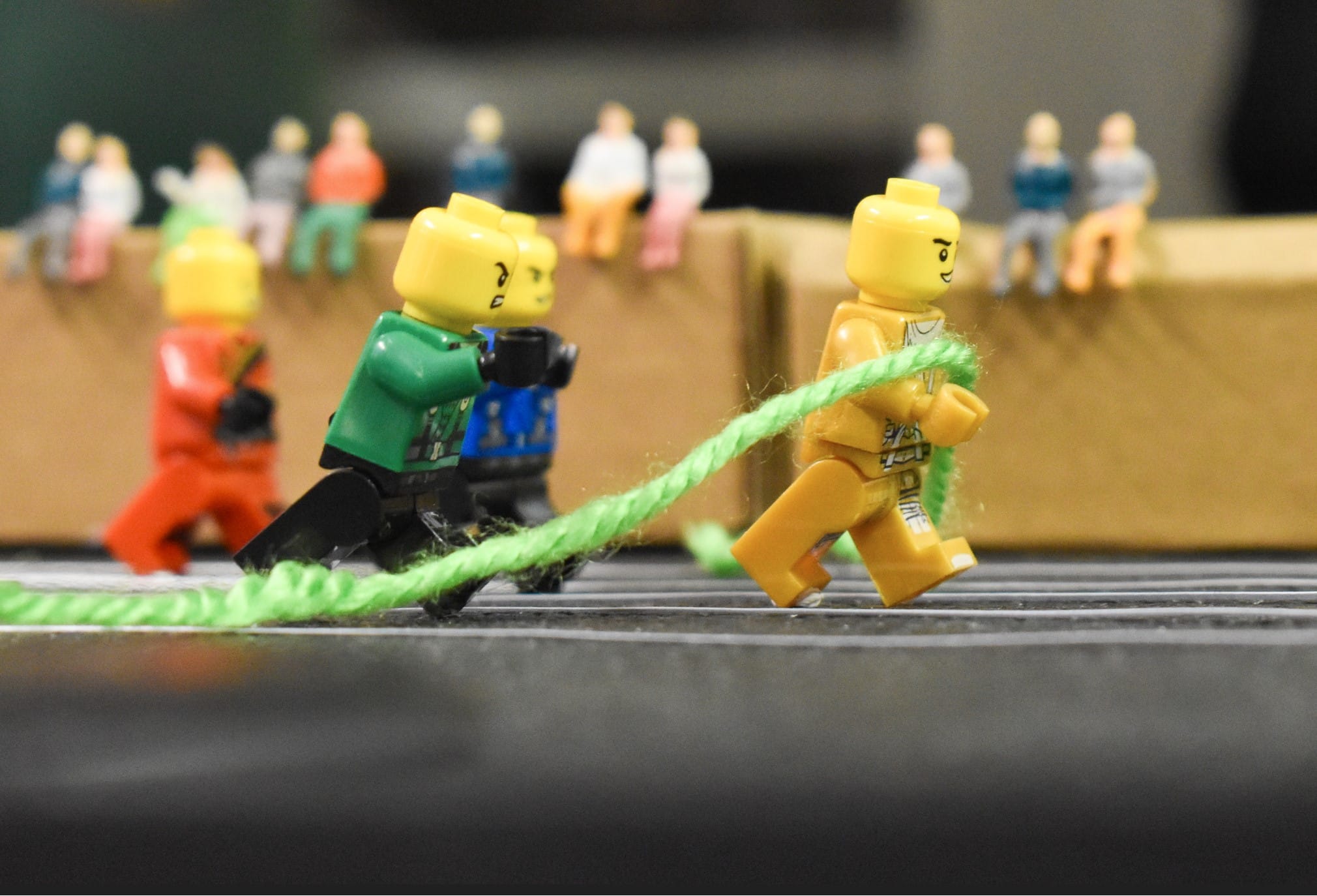 LEGO minifigures playing tug-of-war with a green rope, watched by small spectator figurines in the background.