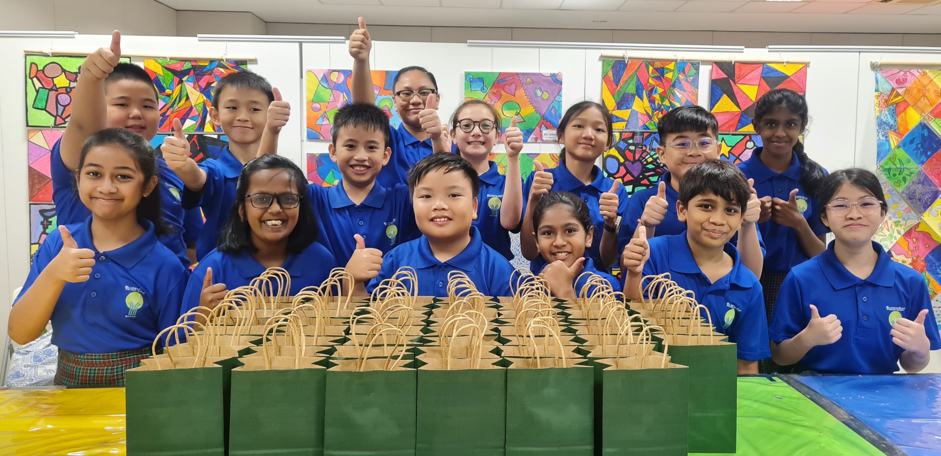 A group of children in blue uniforms giving thumbs up behind rows of green and brown paper bags in a colorful classroom.