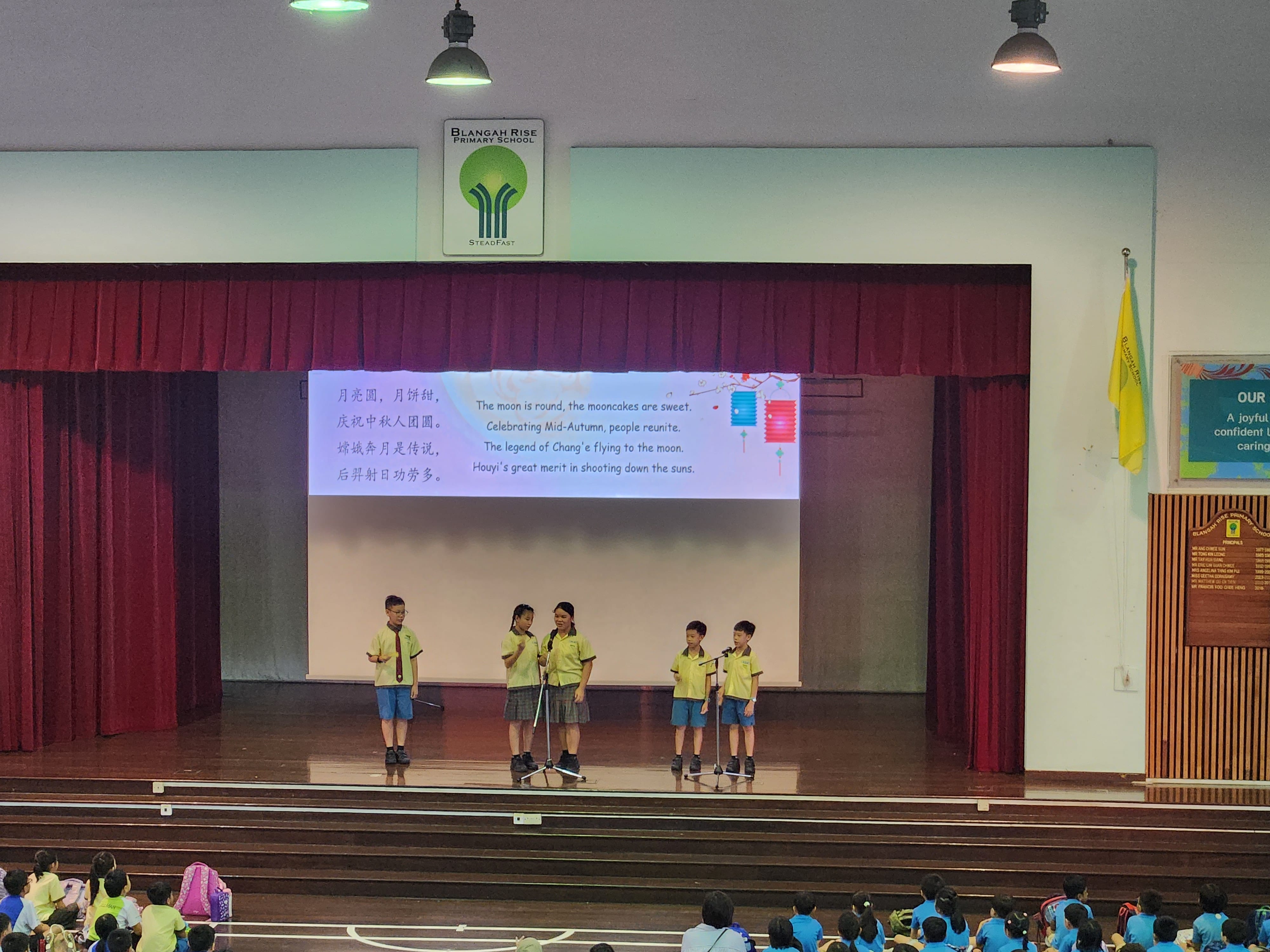 Five students in yellow uniforms recite a Mid-Autumn Festival poem on a school stage with a projected bilingual slide.