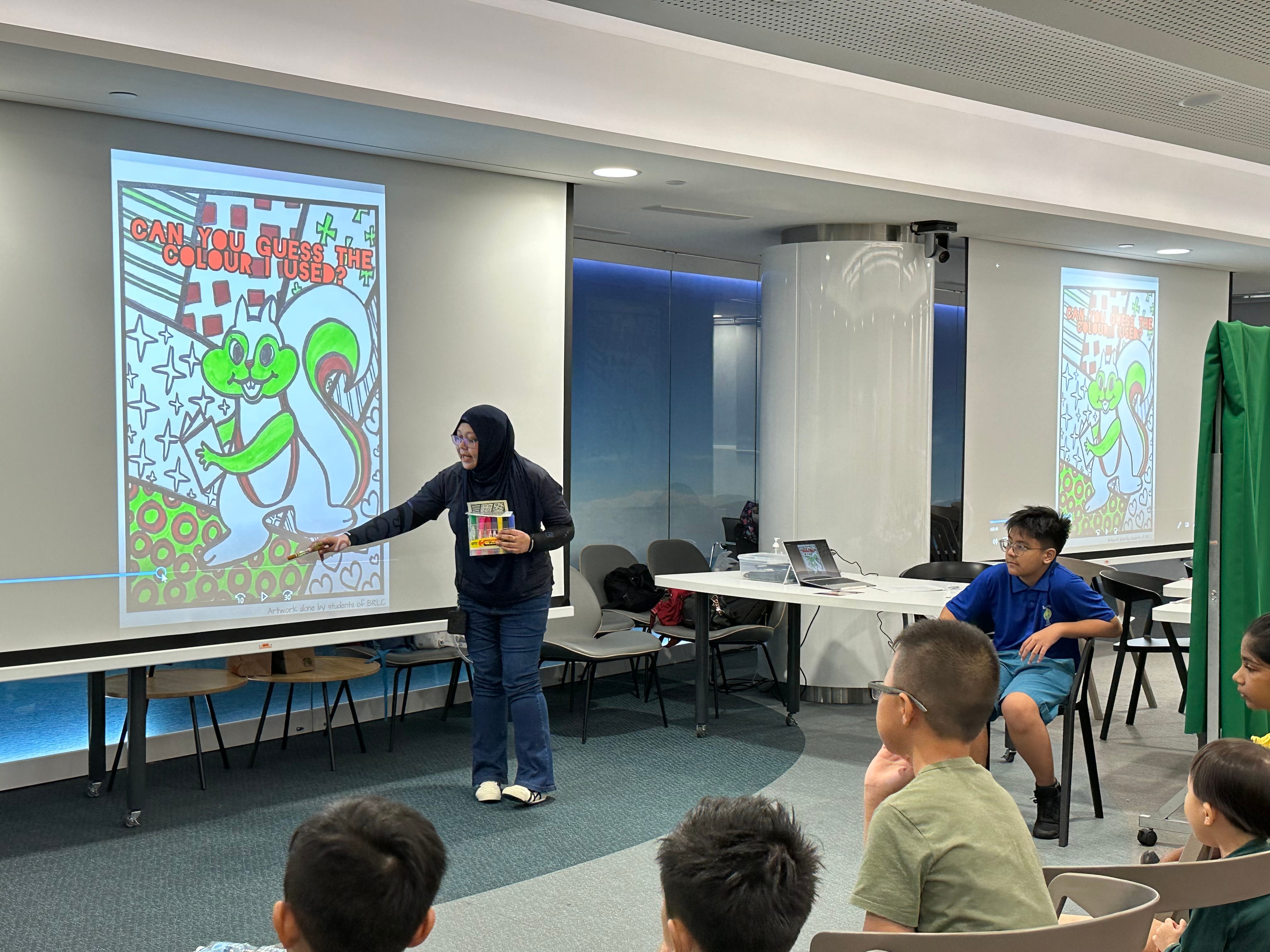 A woman in a hijab points to a projected cartoon squirrel artwork while children watch during an indoor presentation.