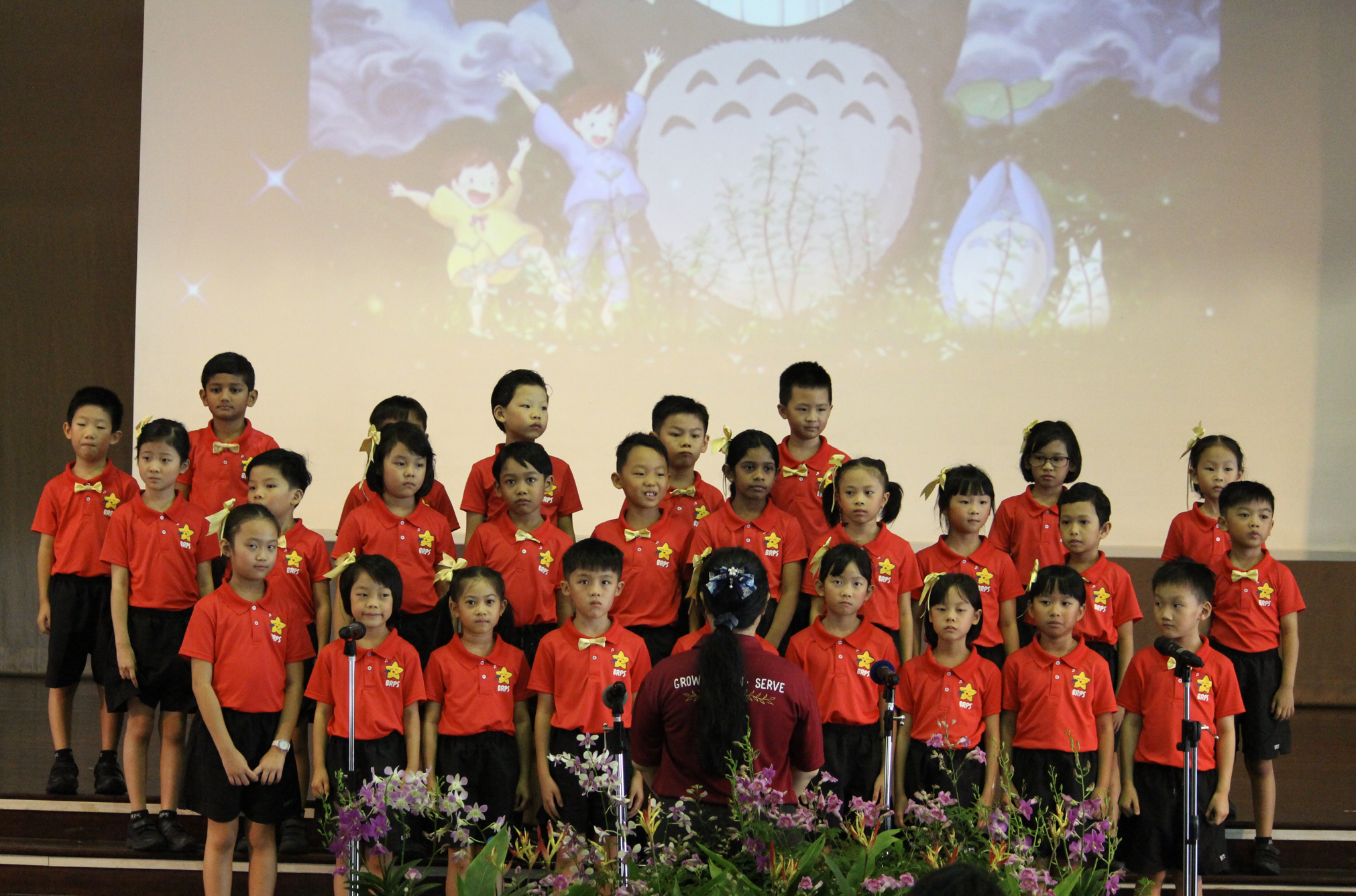 Children in red shirts on stage singing in a choir, conductor in front, cartoon projected behind.