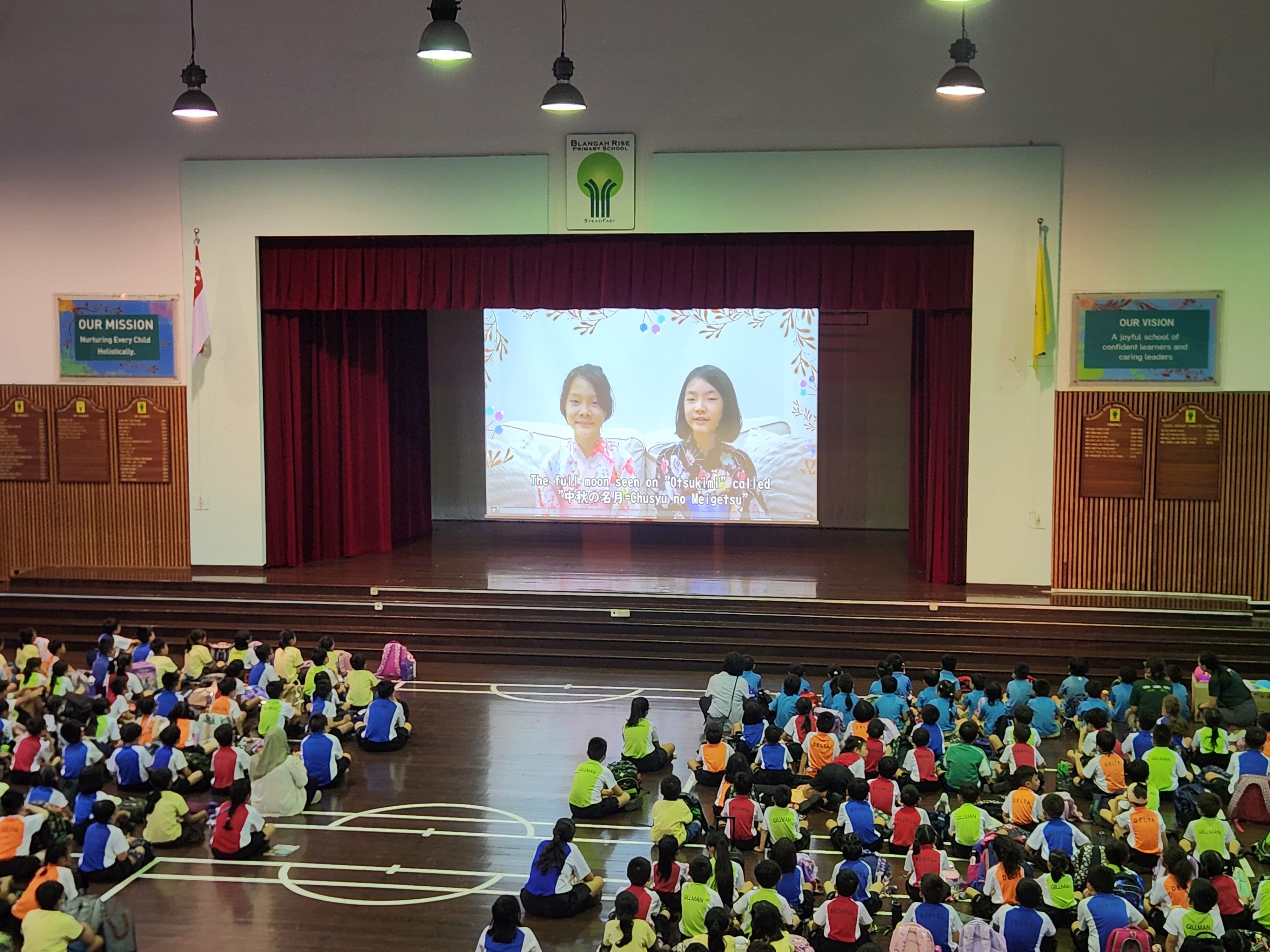 Students seated on gym floor watching a projected video on a stage with red curtains.