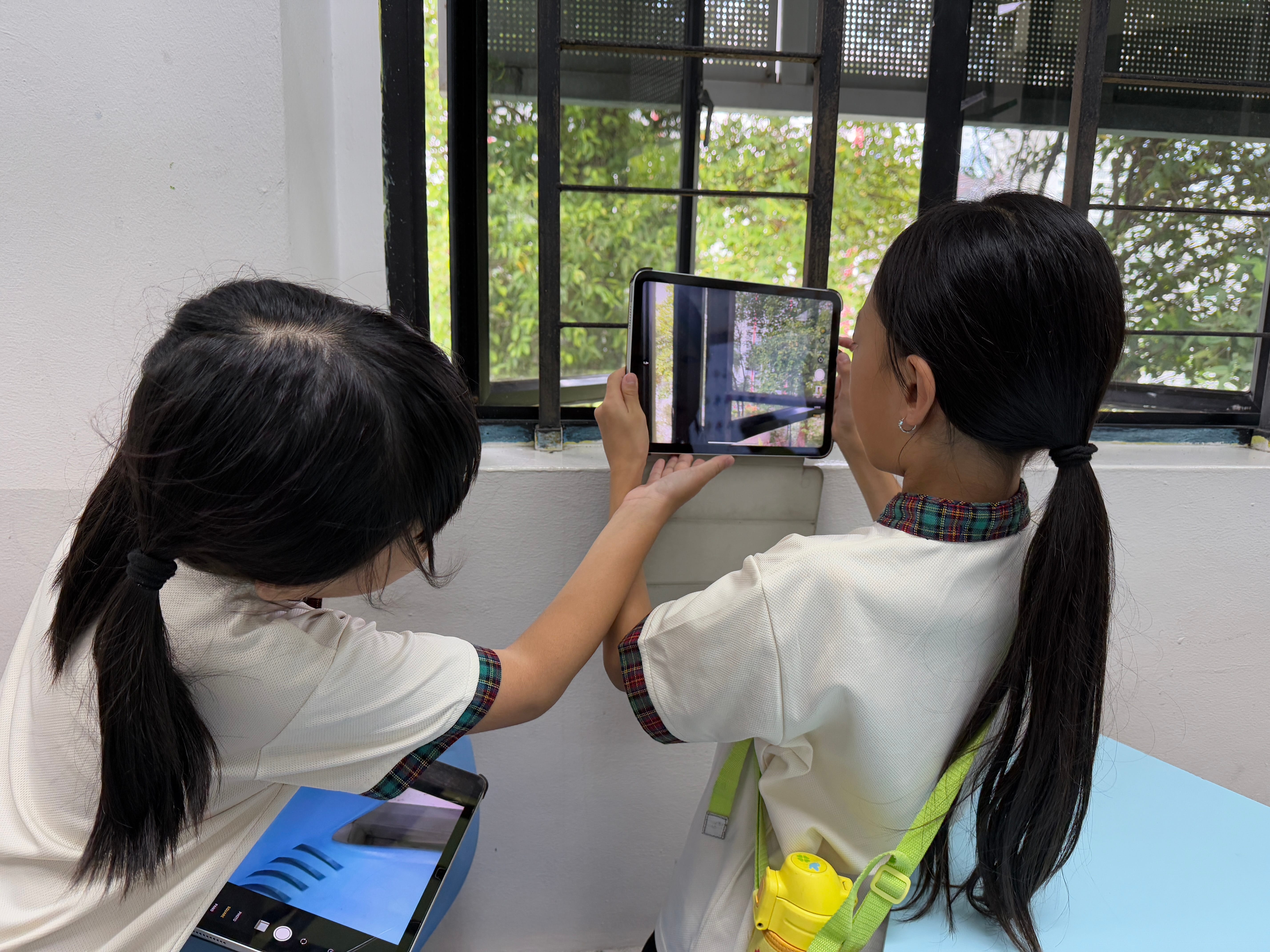 Two girls in white uniforms hold a tablet up to a barred window, photographing trees outside.