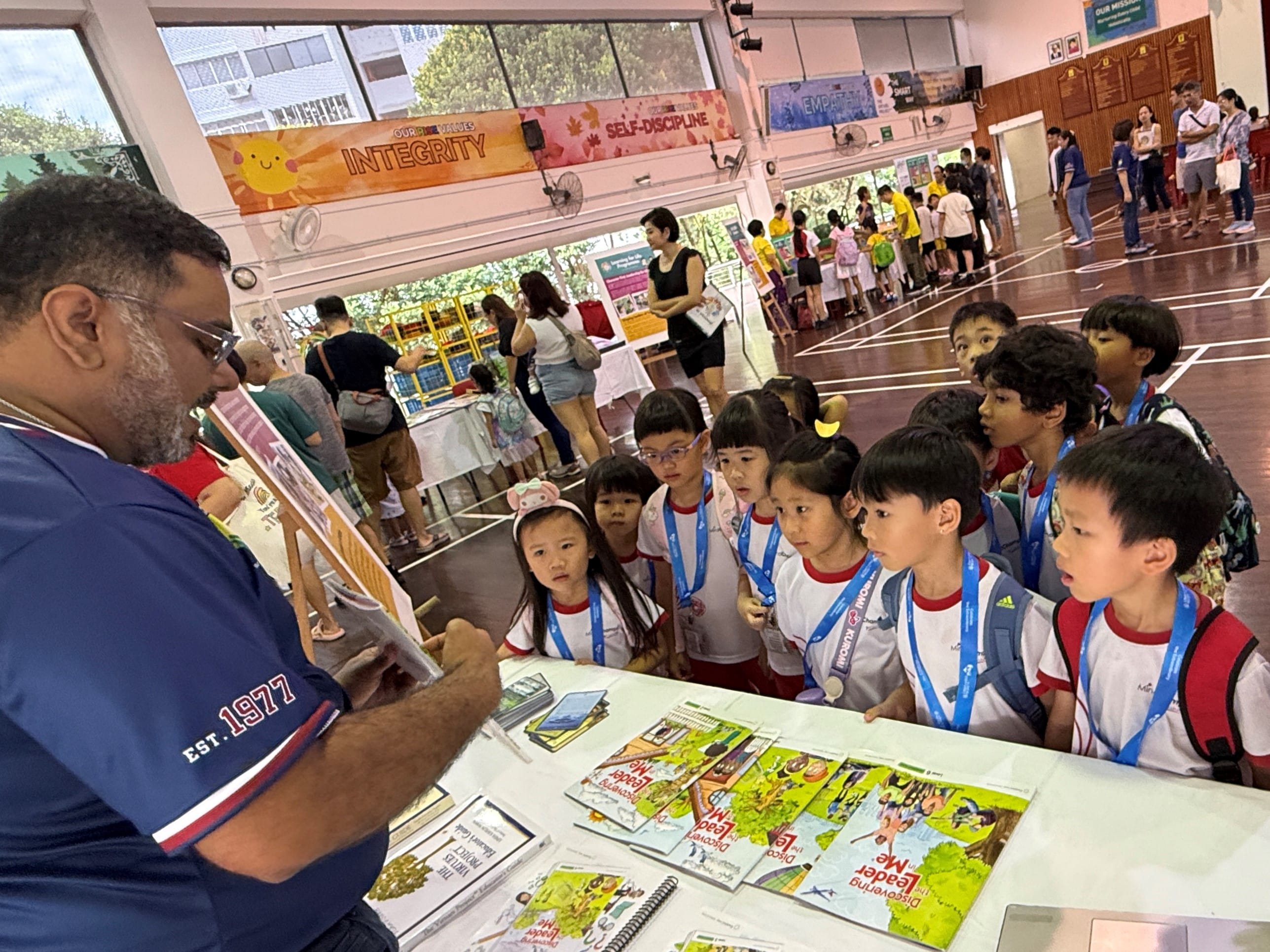 Man at a table shows books to schoolchildren with medals around their necks in a busy indoor hall.