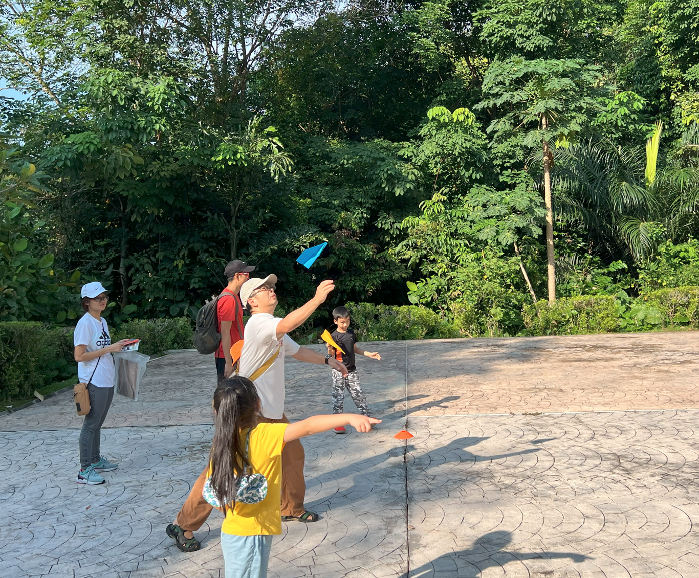 A group of adults and children throwing colorful paper airplanes on an outdoor paved area surrounded by green trees.