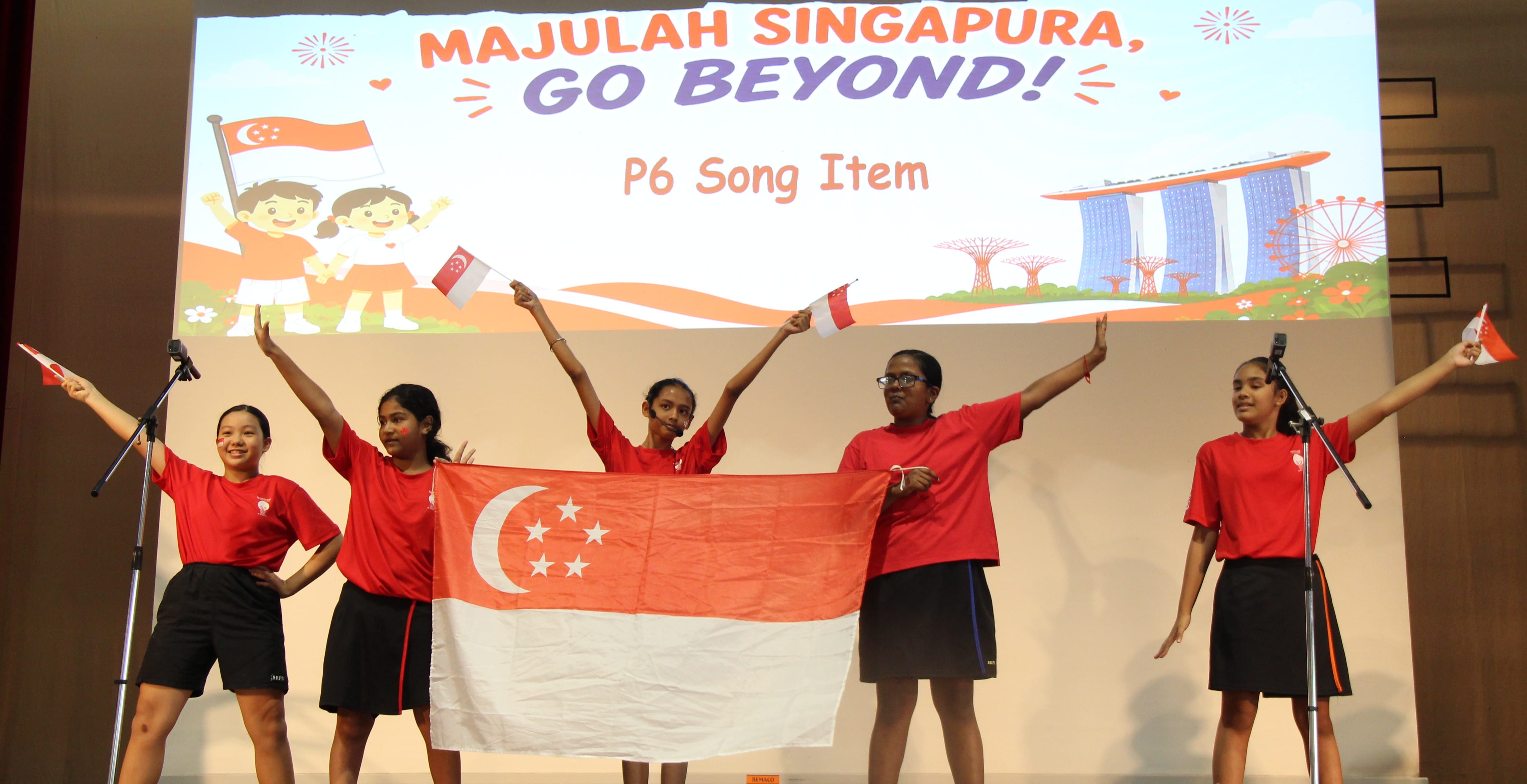 Five students in red shirts on stage hold a Singapore flag under a "Majulah Singapura, Go Beyond! P6 Song Item" banner.