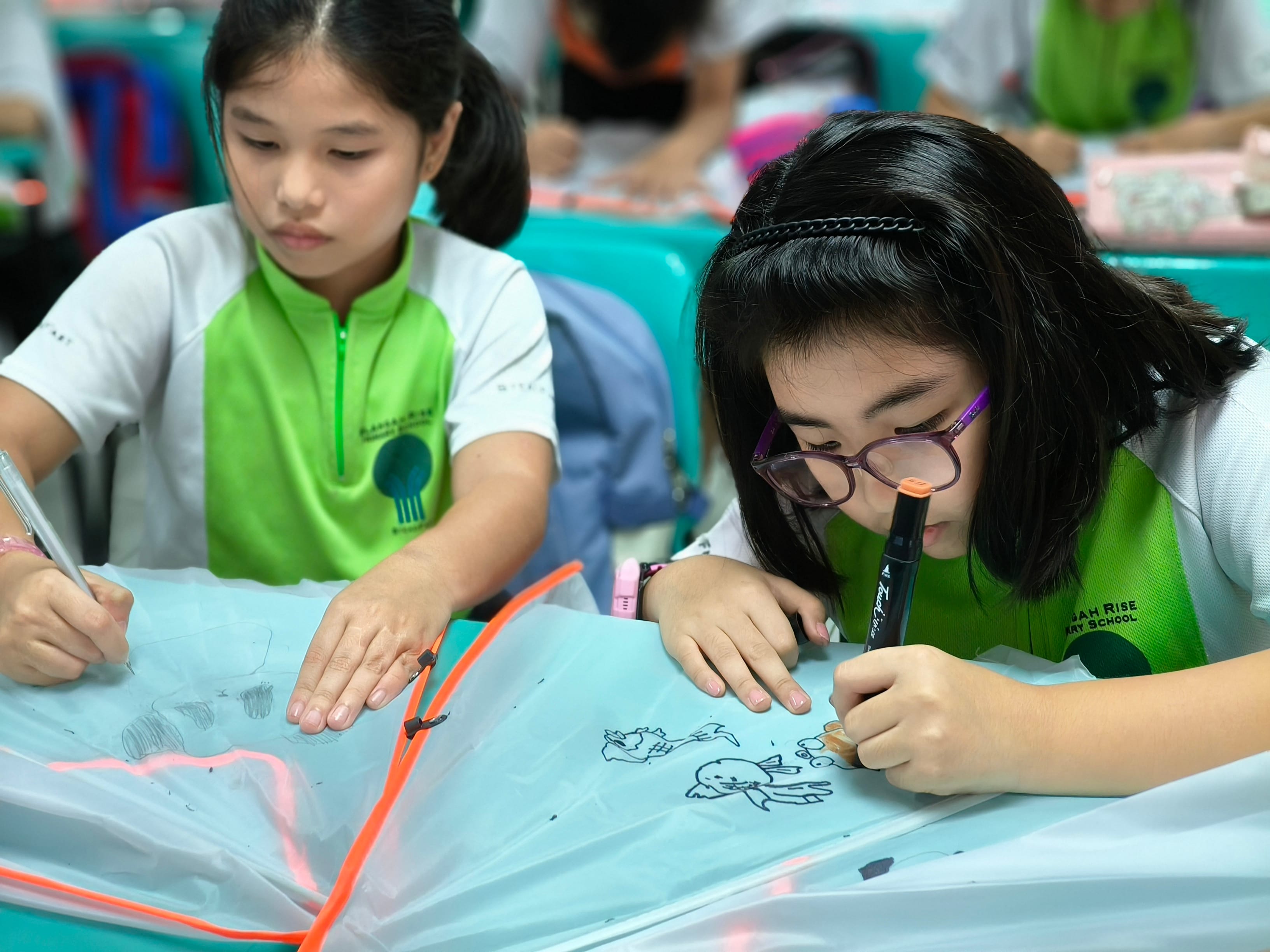 Two schoolgirls in green uniforms sit at desks, drawing on clear plastic sheets with pens.