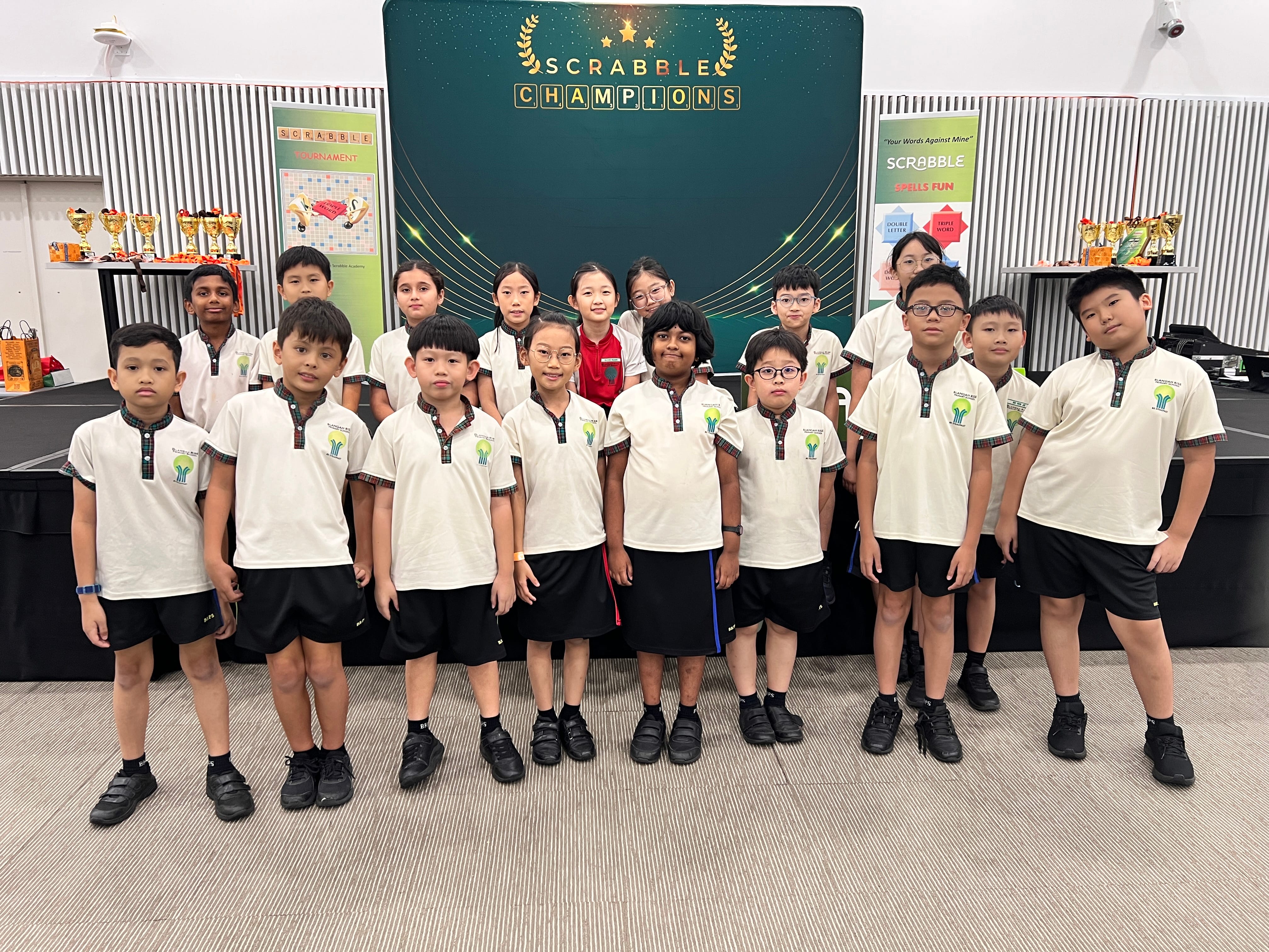 Group of schoolchildren in uniform posing before a Scrabble Champions backdrop with trophies on tables.
