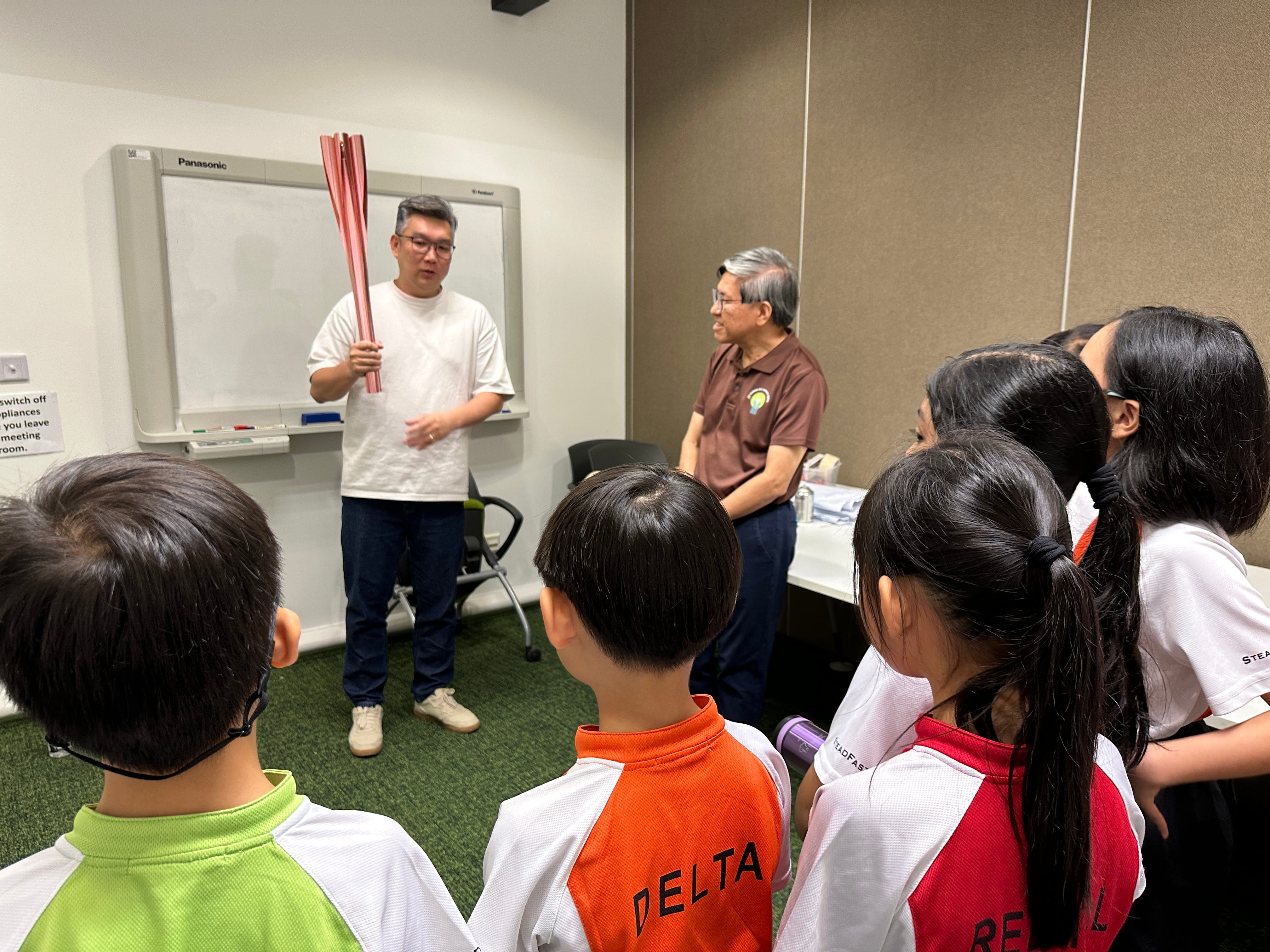 A man holds pink tubes while presenting to a group of children in school uniforms inside a meeting room.